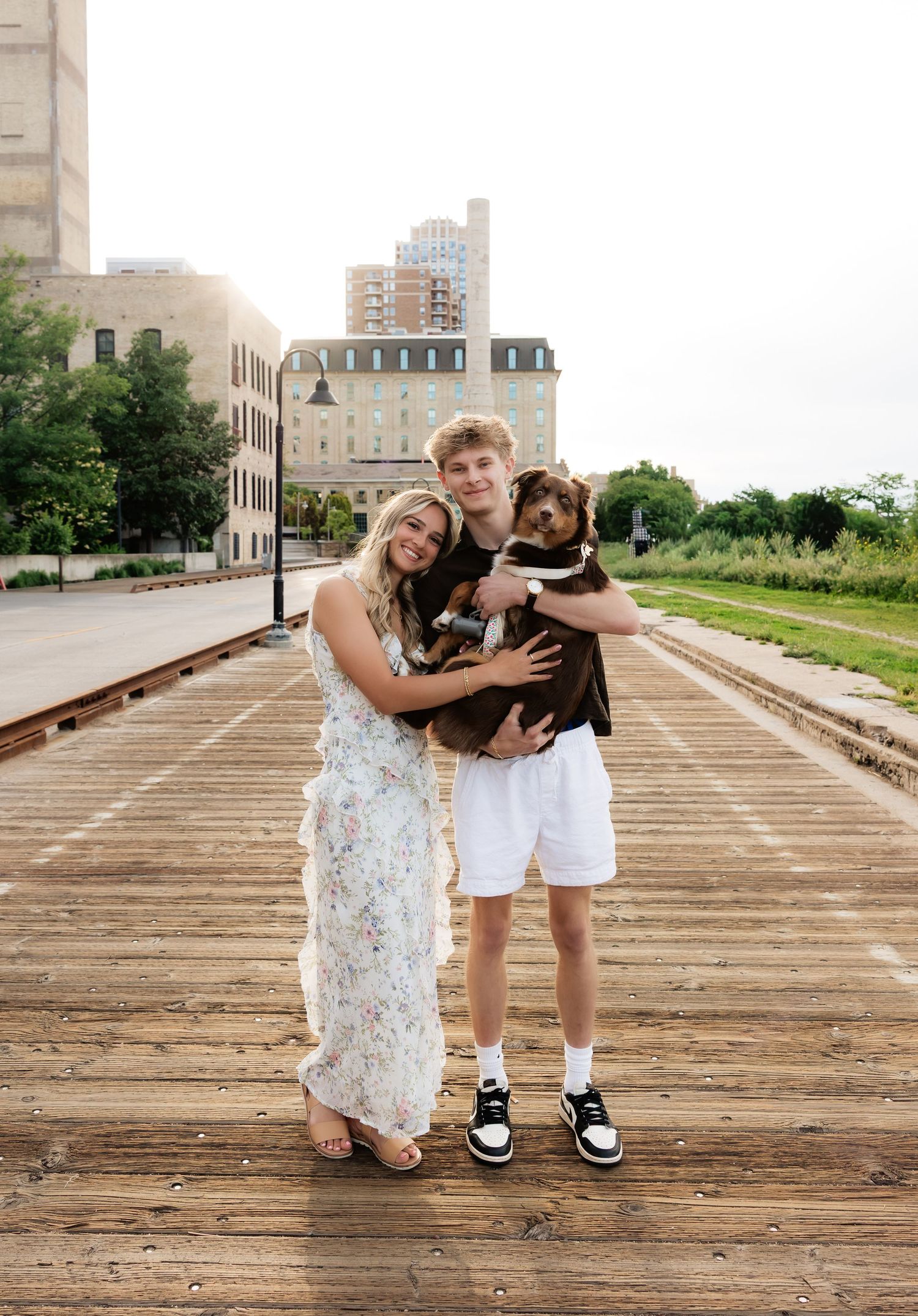 Young couple smiling and holding their brown Australian Shepherd dog on a wooden boardwalk in an urban setting.