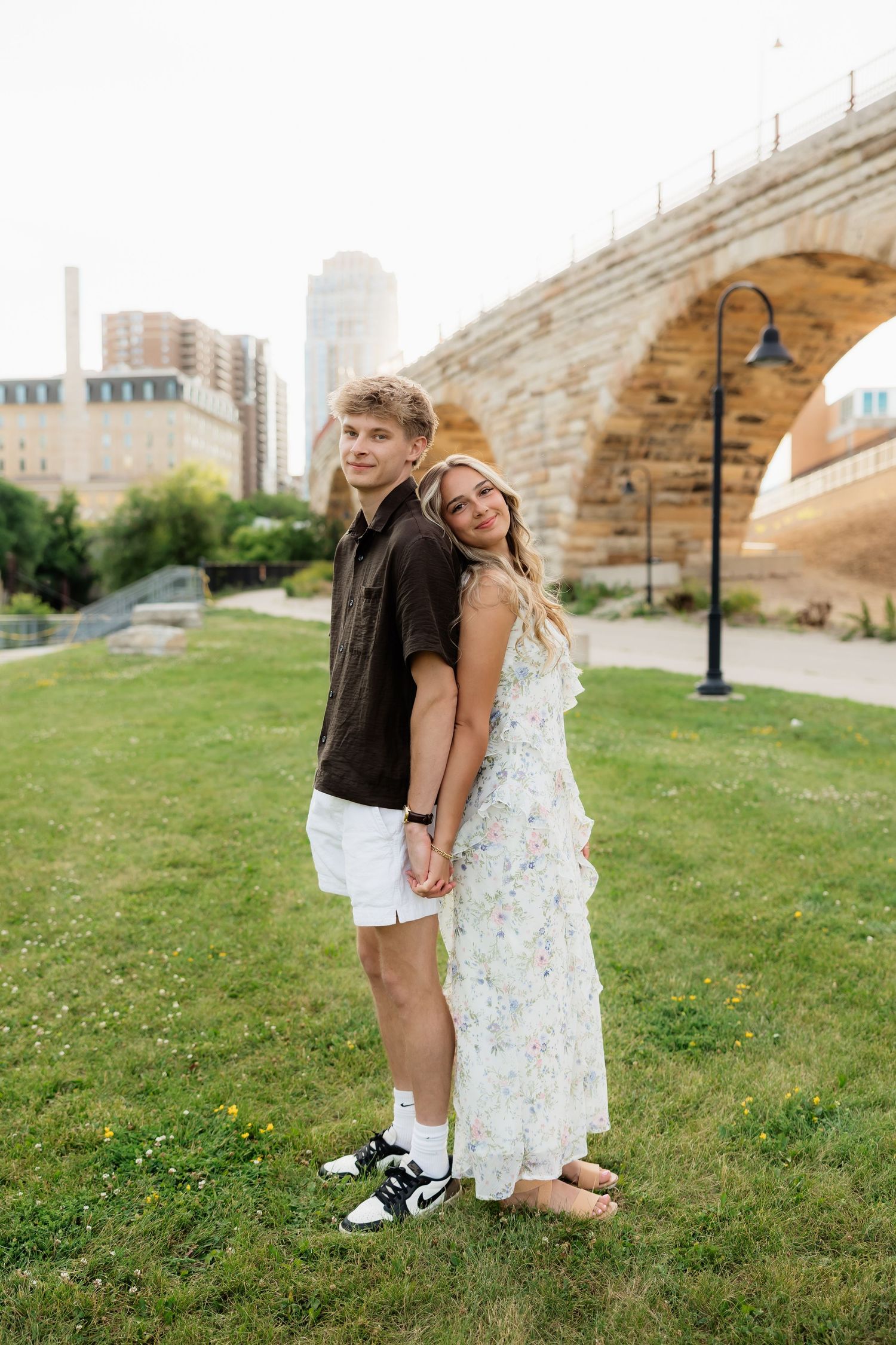 Couple posing back-to-back on green grass near a stone arch bridge, woman in floral dress leaning on man.