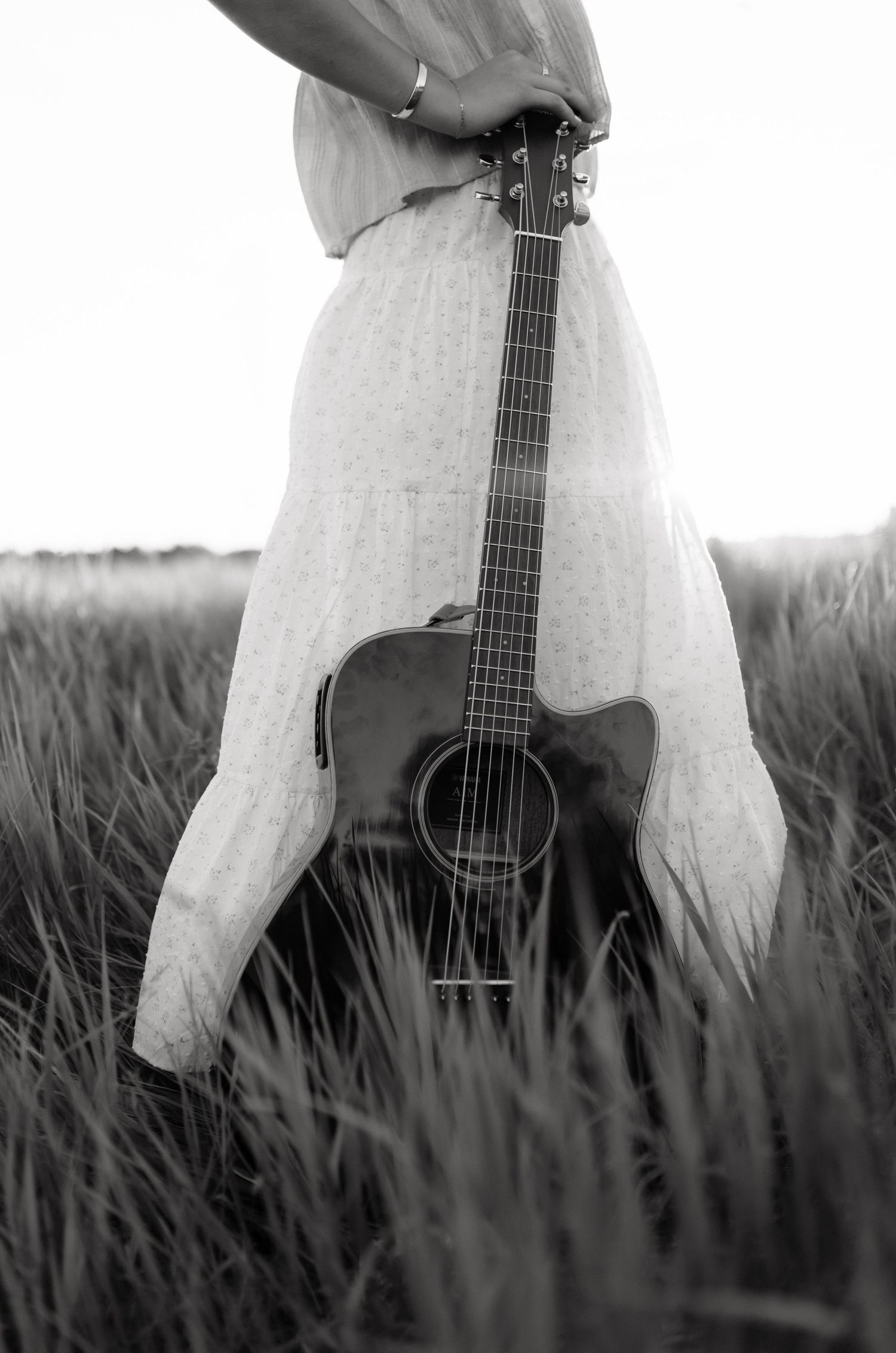 Black and white photo of woman in white dress holding acoustic guitar in tall grass field.
