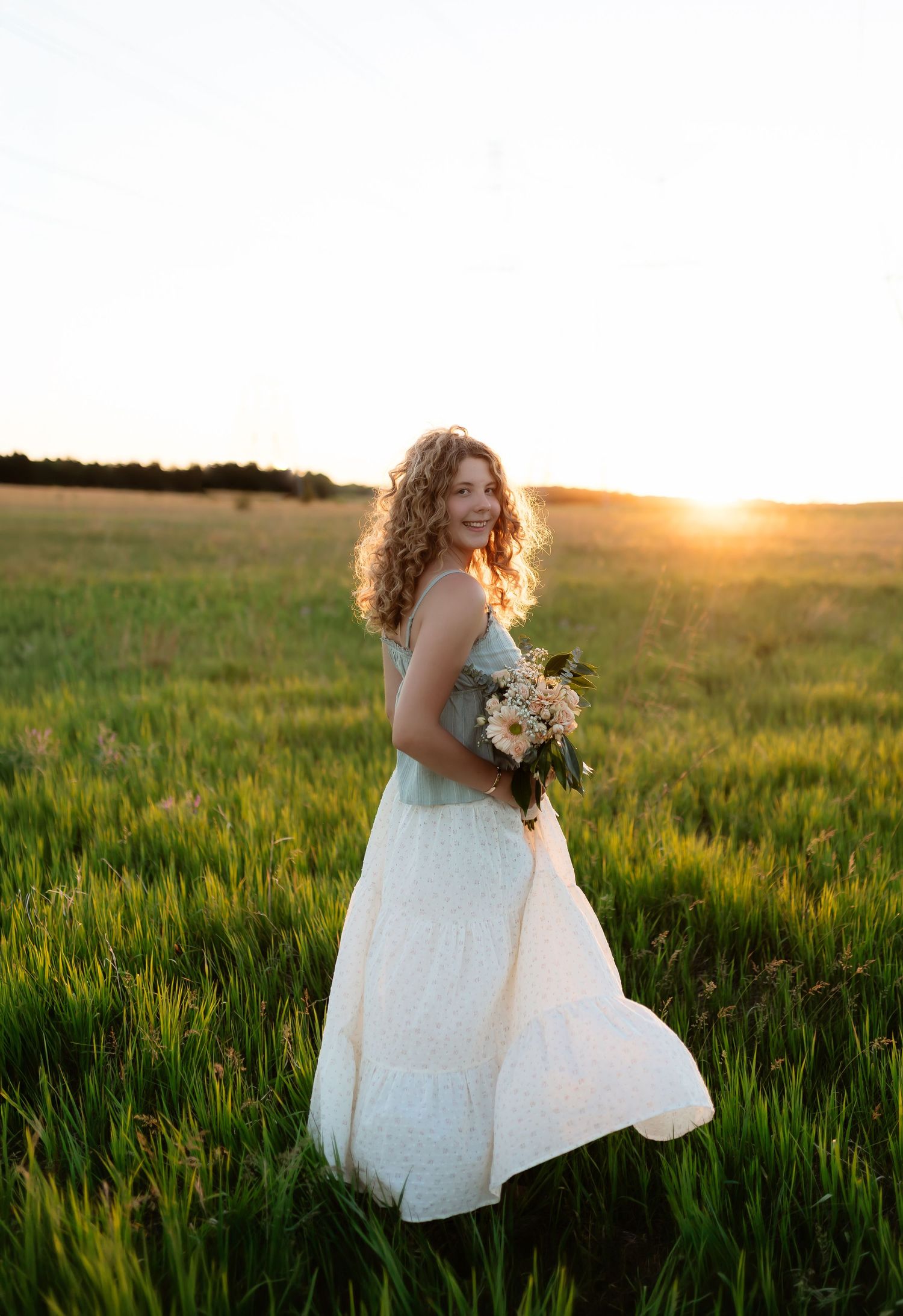Smiling young woman with curly hair holding a flower bouquet in a green field bathed in warm golden sunset light.