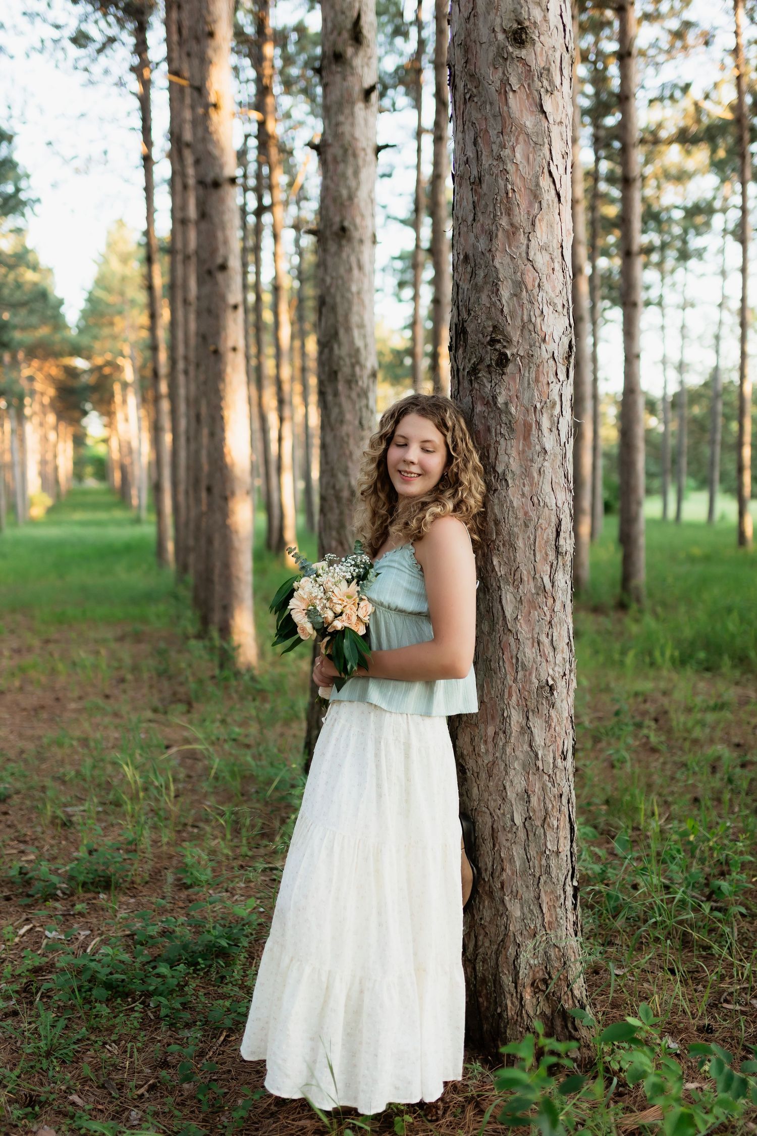 Smiling young woman in mint and white dress holding a bouquet while leaning against a pine tree.