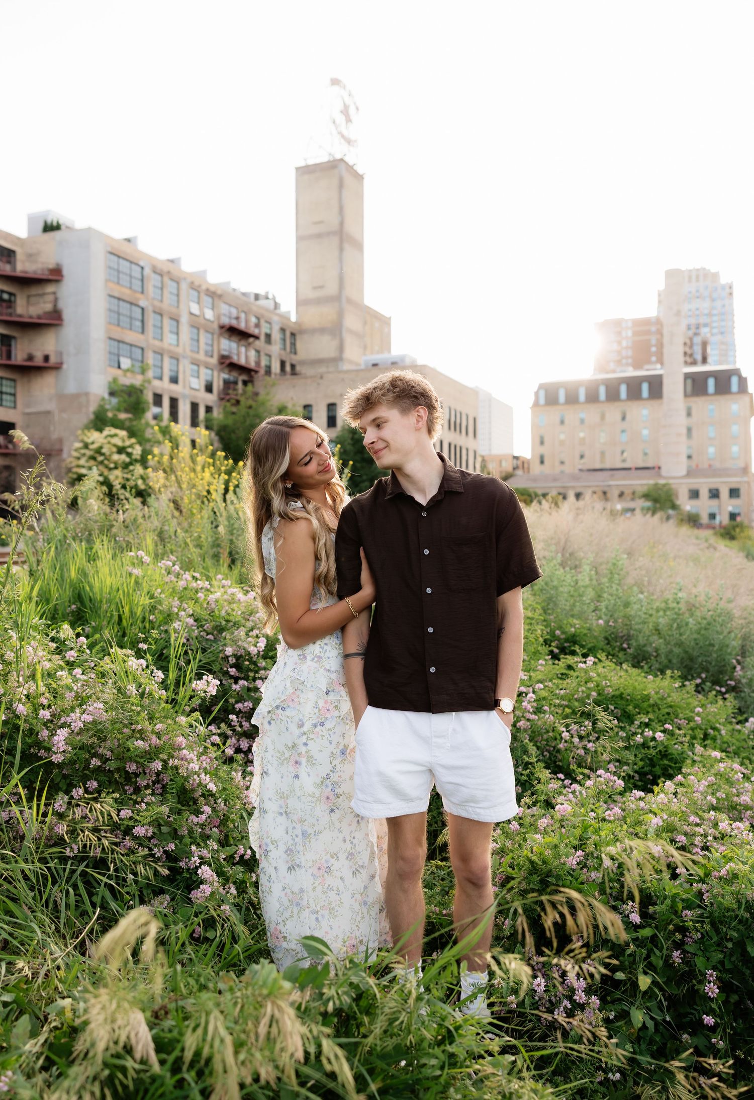 Young couple standing among blooming wildflowers in urban garden, woman gazes at tall man in black shirt.