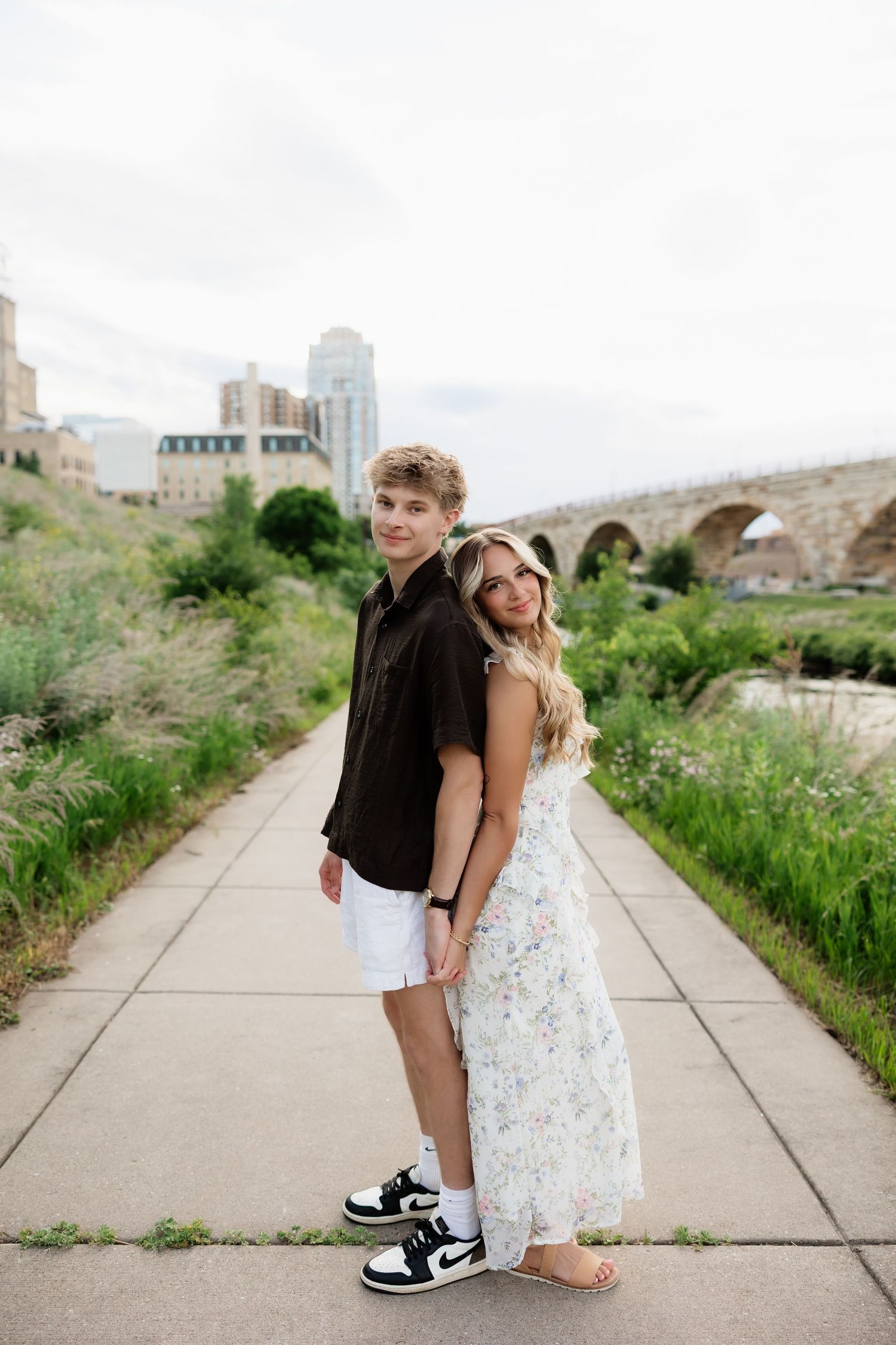 Young couple posing back-to-back on urban pathway, stone bridge behind them, man in black shirt and sneakers.