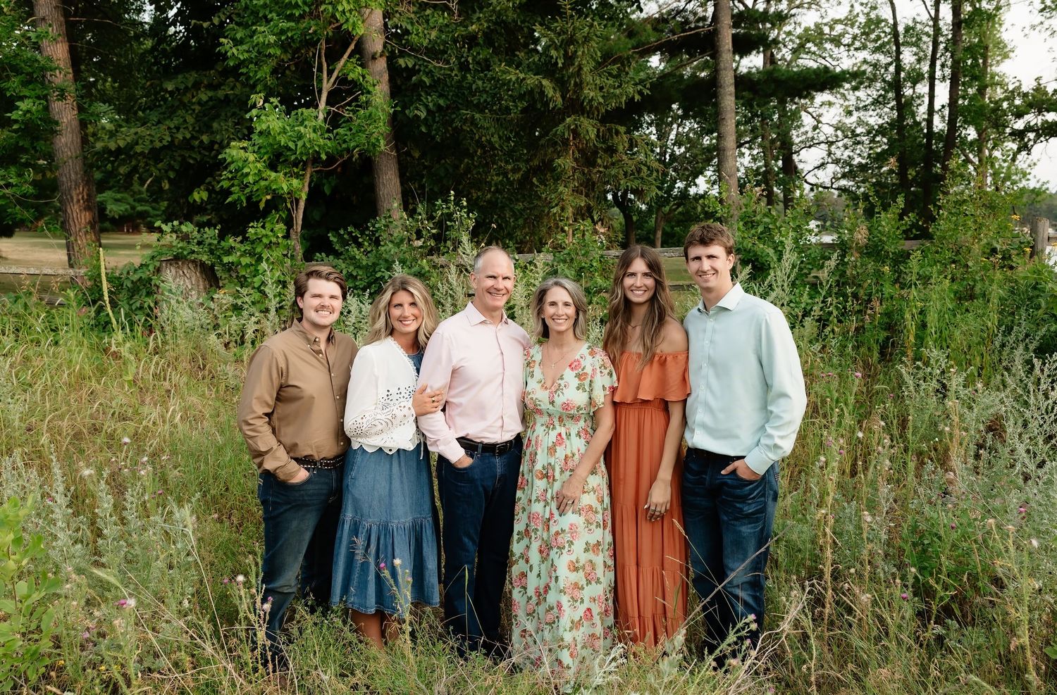 A happy family of six poses together in a lush green outdoor setting, wearing coordinated pastel outfits.