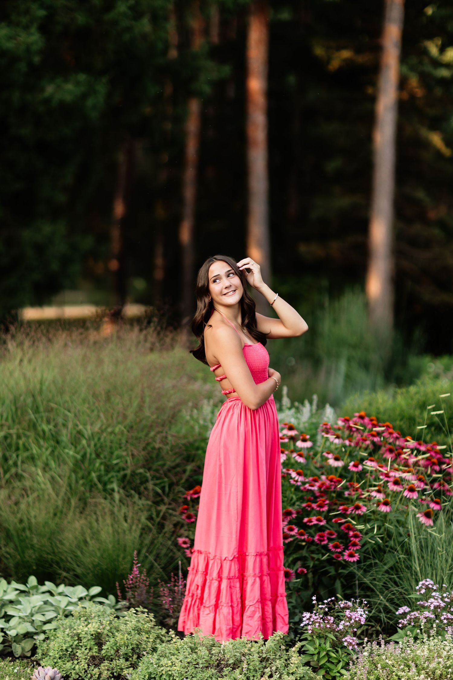 Young woman in pink maxi dress posing among colorful garden flowers with lush green forest background.