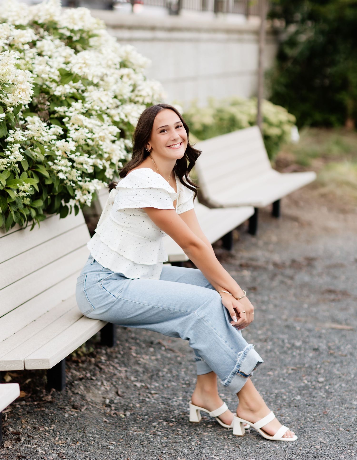 Smiling young woman in white off-shoulder top and light jeans poses on bench near white hydrangeas.