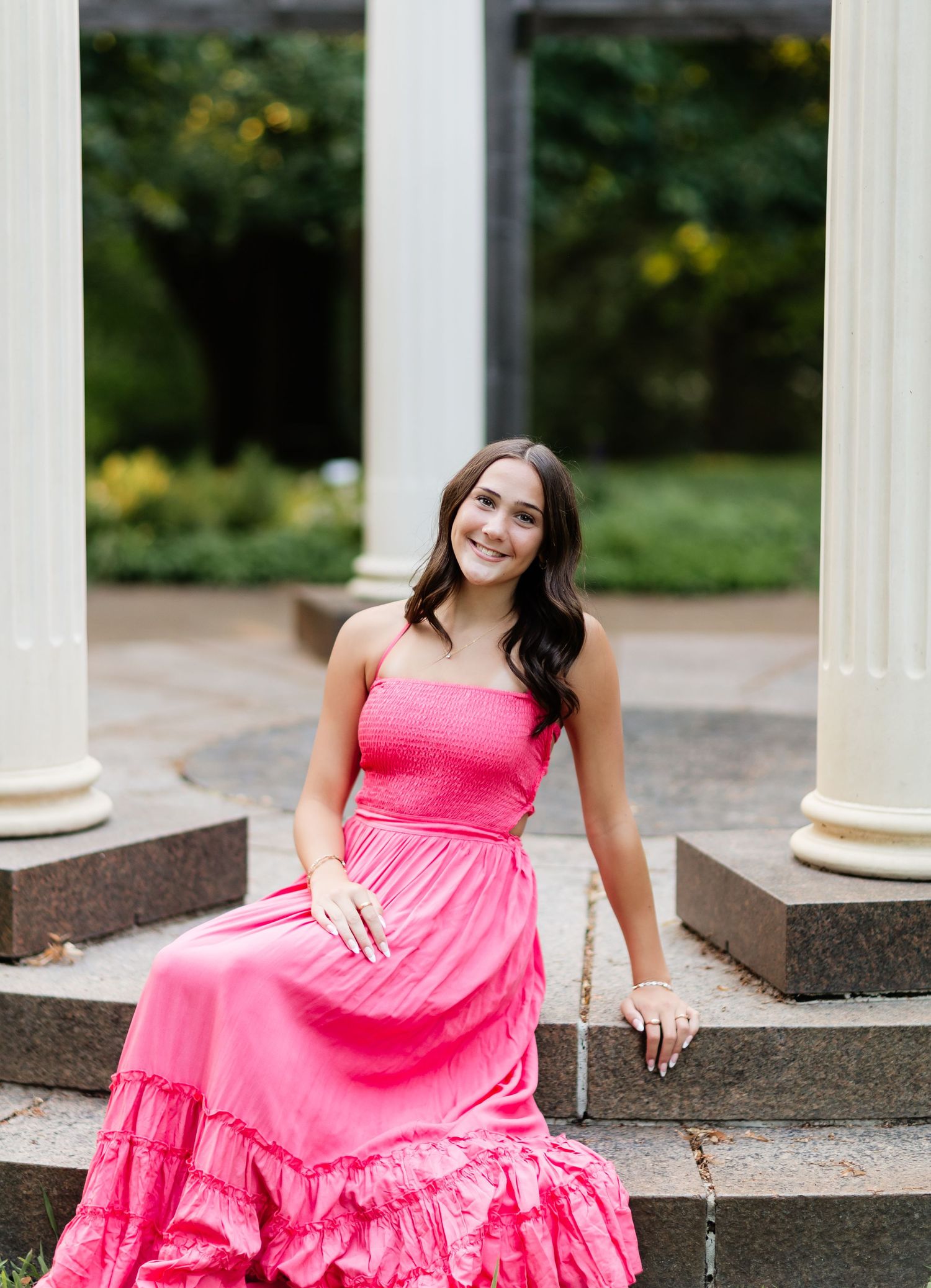 Smiling young woman in a pink strapless maxi dress seated on stone steps between white columns.
