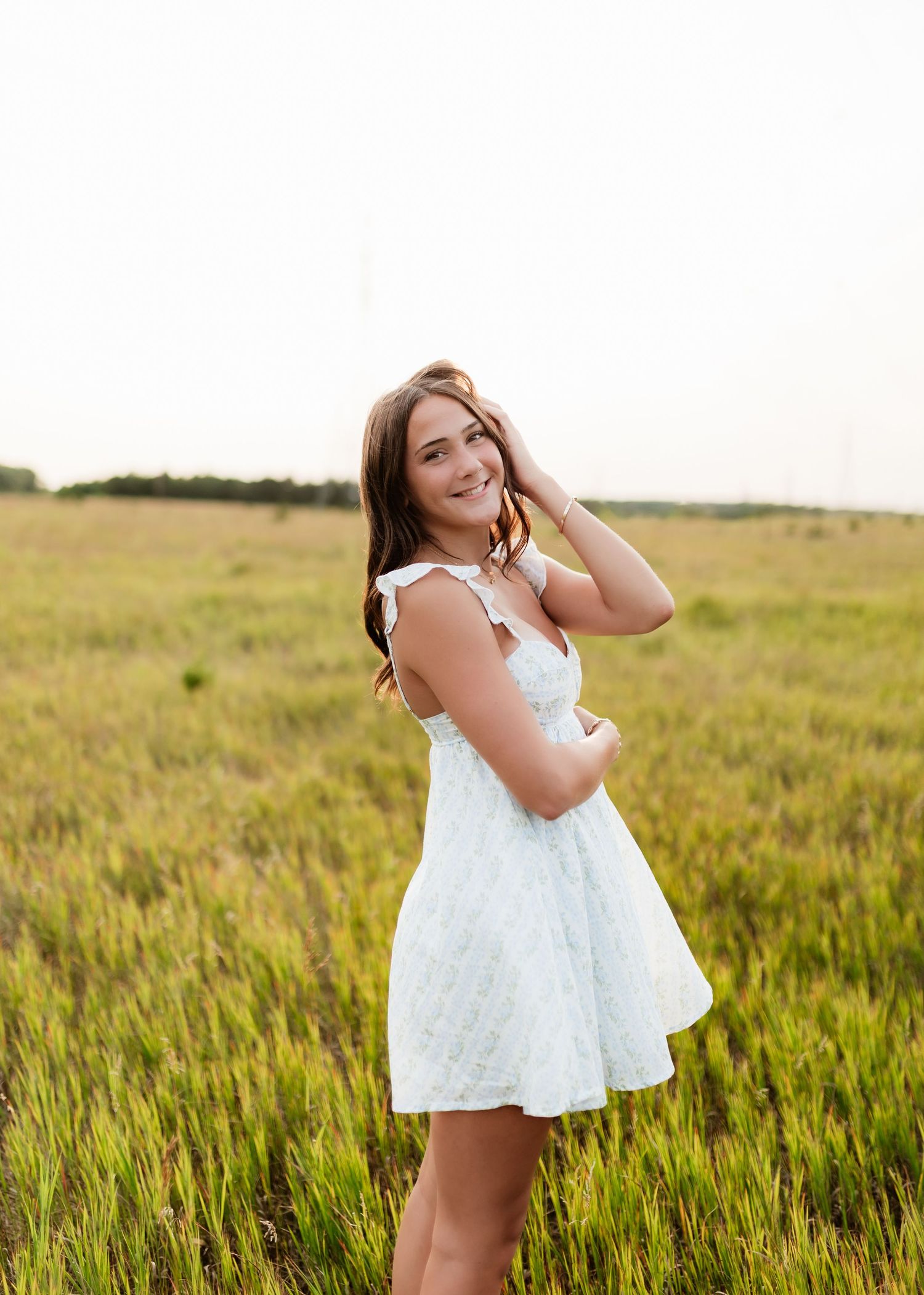Smiling brunette in white floral dress posing in lush green meadow at dusk.