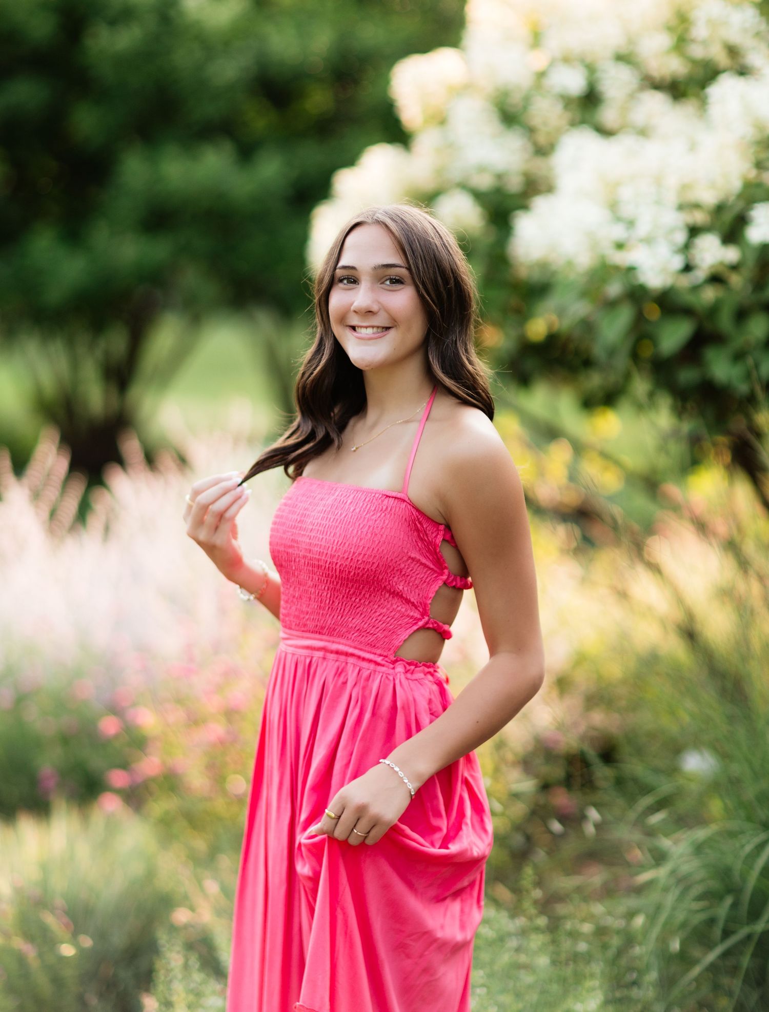 Smiling young woman in a vibrant pink halter sundress stands among lush garden blooms and greenery.