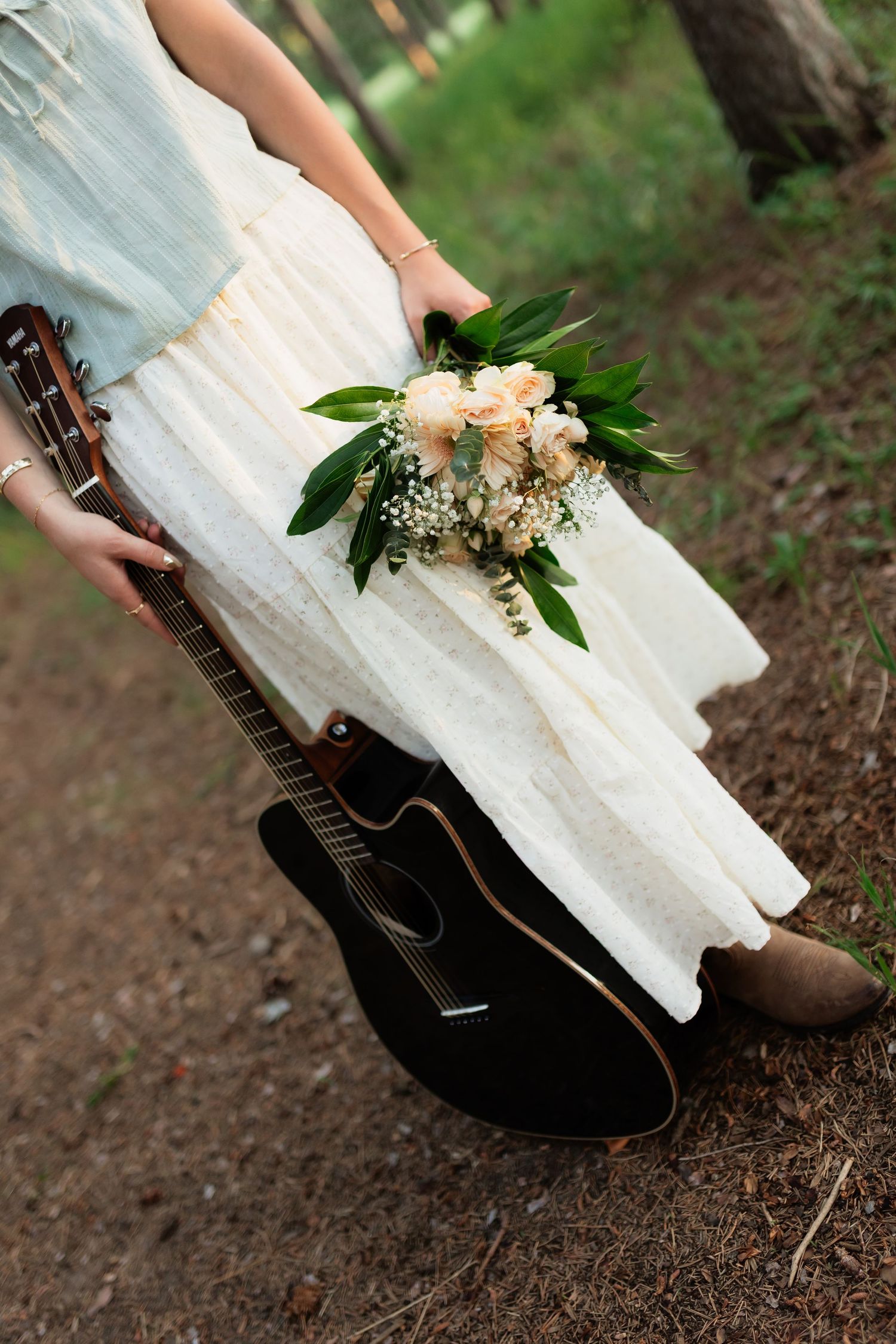 Woman in white dress holding floral bouquet beside black acoustic guitar outdoors in forest setting.