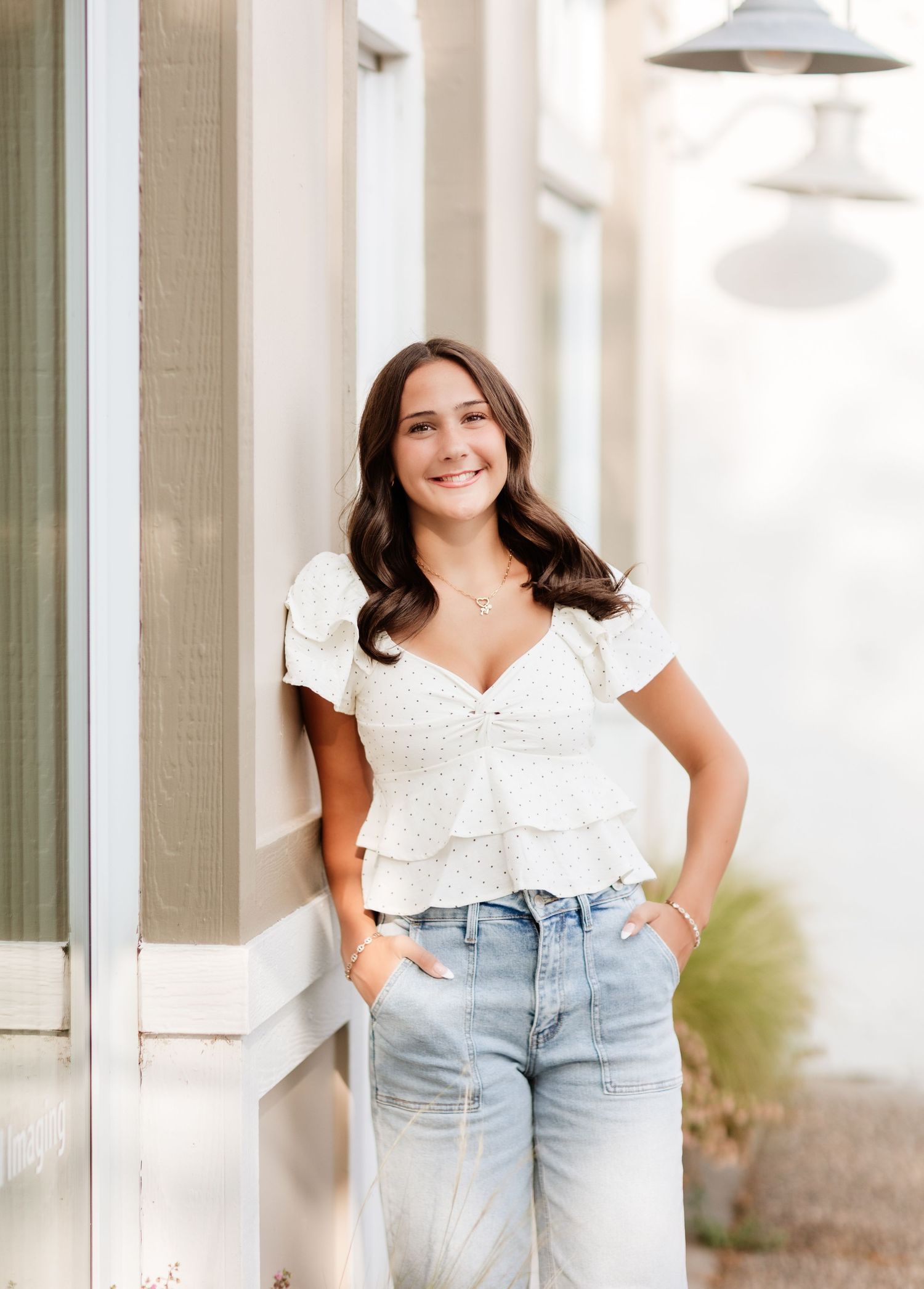 Smiling brunette teen in white eyelet ruffle top and light wash jeans leaning against a wall.