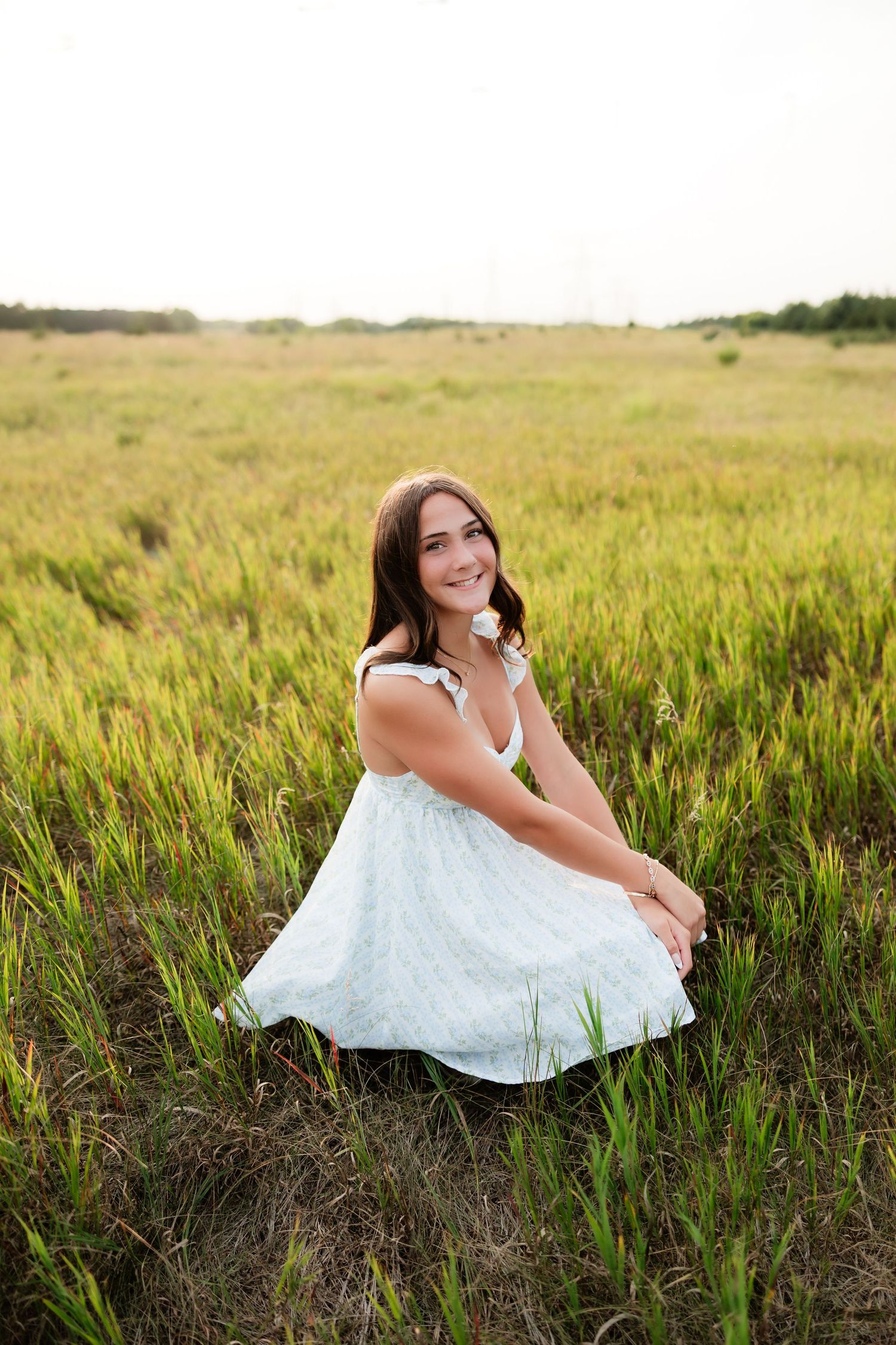 Young woman in white sundress sitting in green field, smiling, holding white flower outdoors.