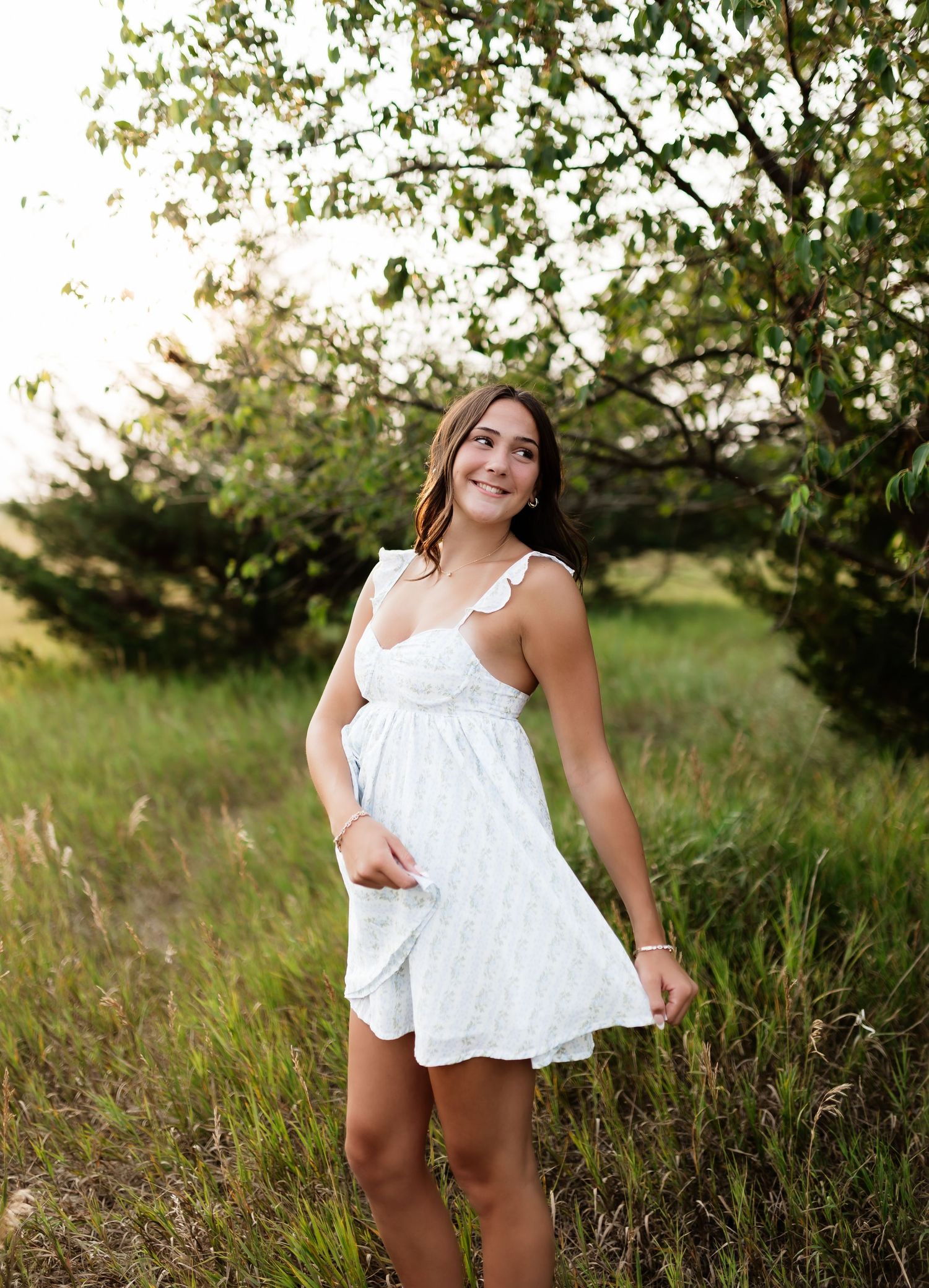 Young woman in white sundress standing in green meadow near trees, smiling in golden hour light.