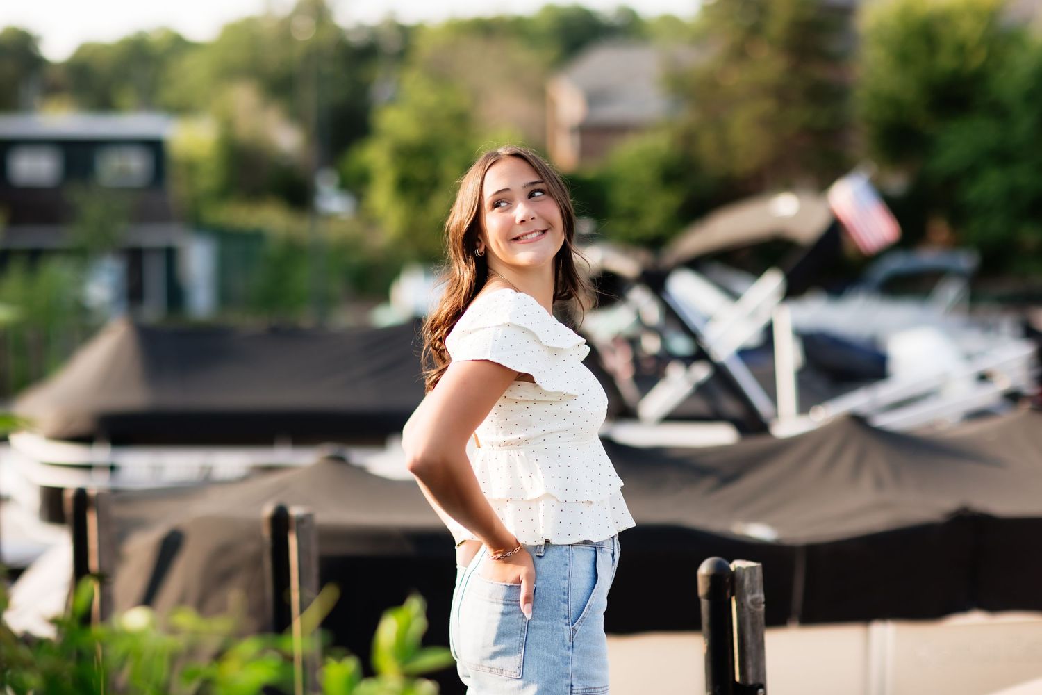 Young woman in white off-shoulder top and jeans smiling confidently at a marina with boats in background.