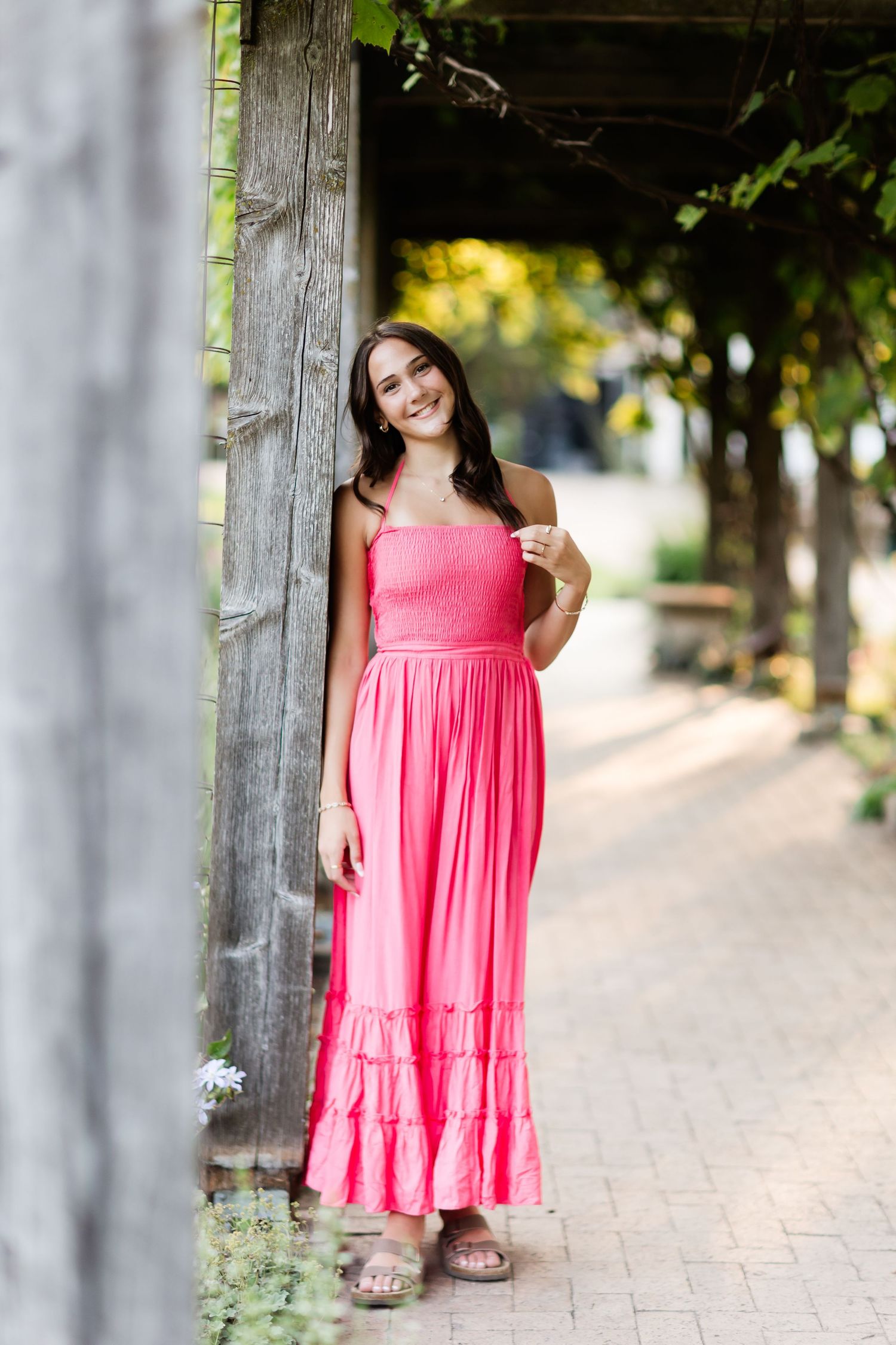 Smiling young woman in pink ruffle maxi dress leaning against rustic wooden pergola post outdoors.