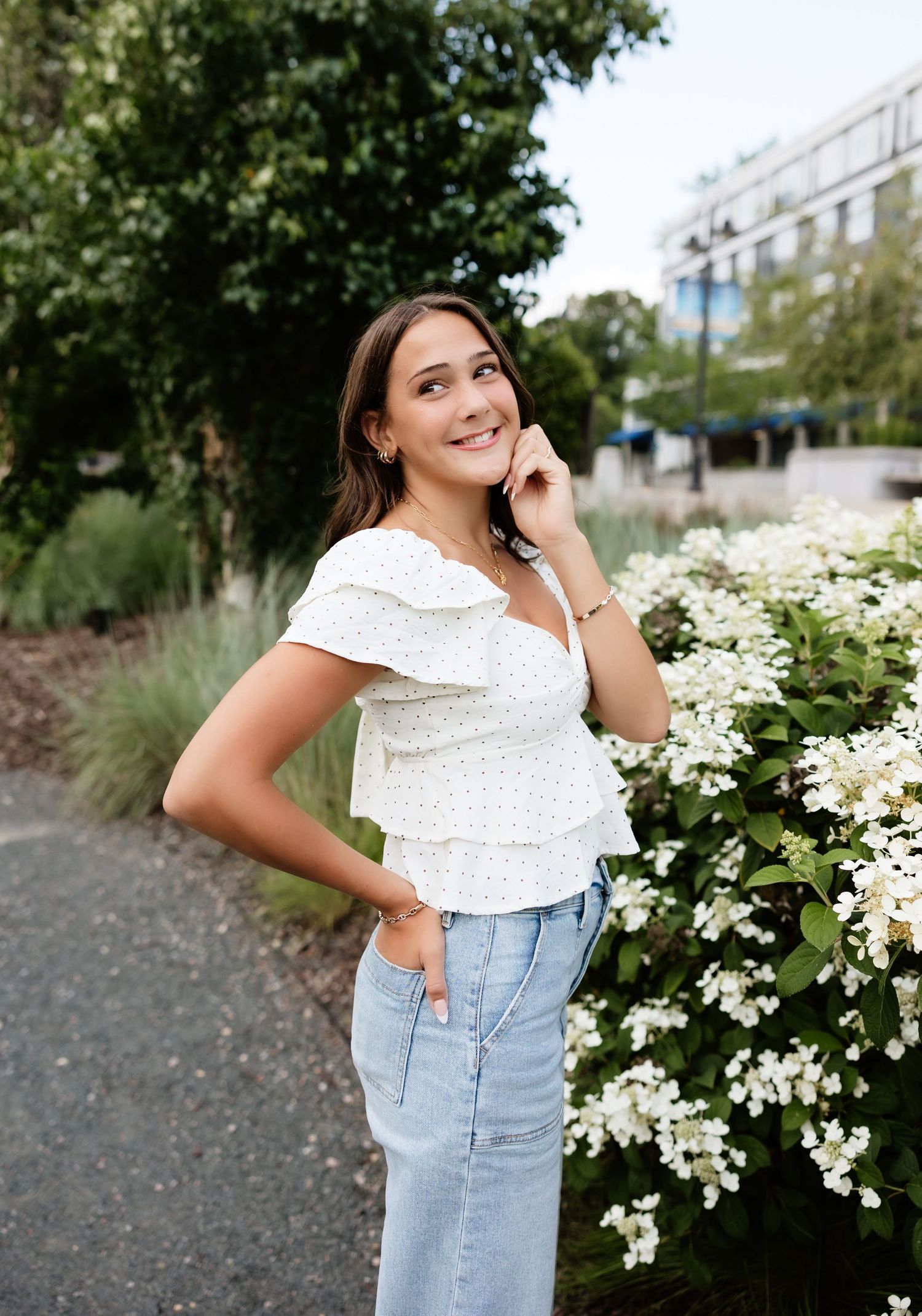 Young woman in white ruffled top and jeans smiling outdoors near white flowering bushes.