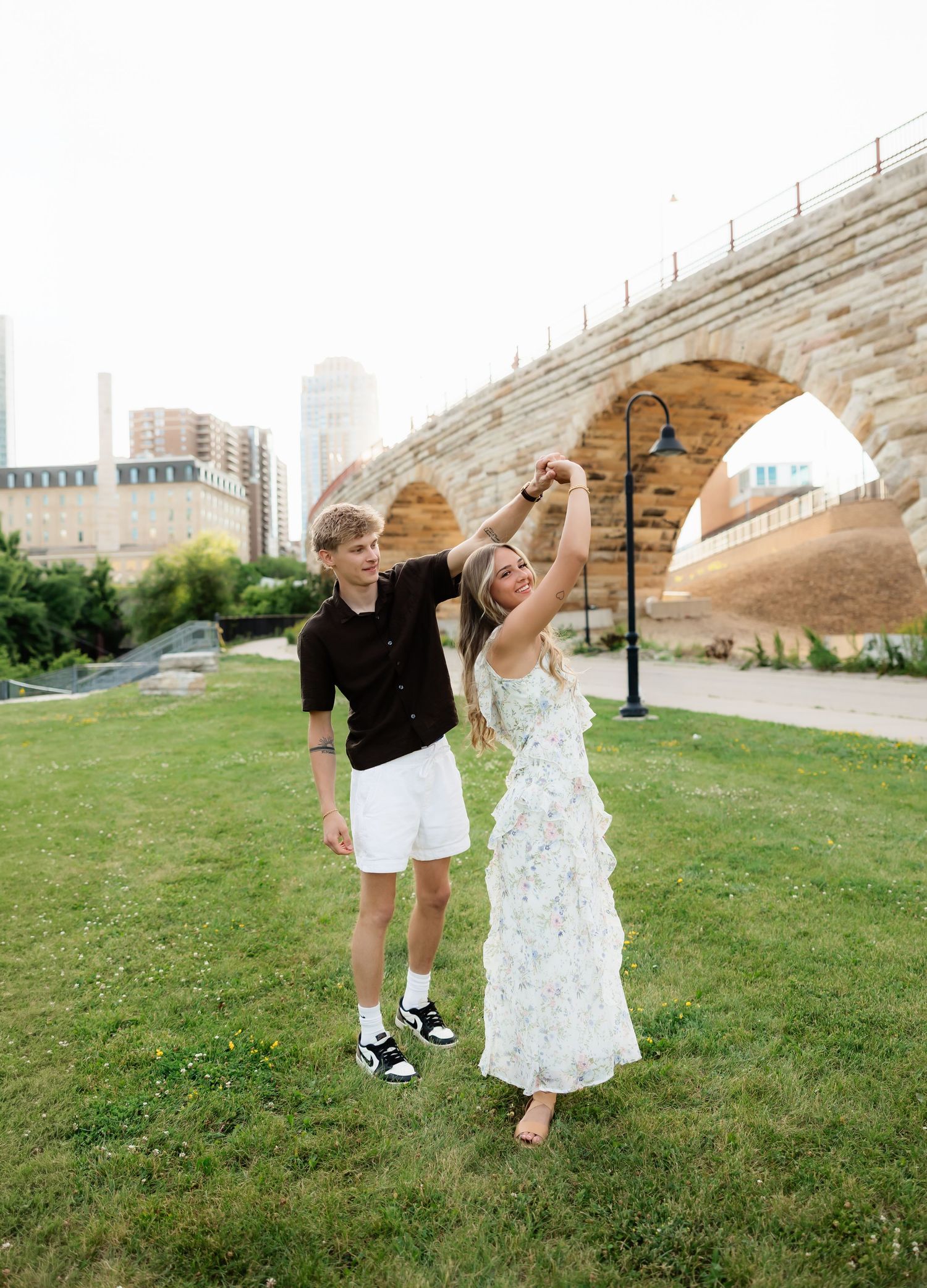 Couple dancing on green grass with a historic stone arch bridge and city skyline in the background.