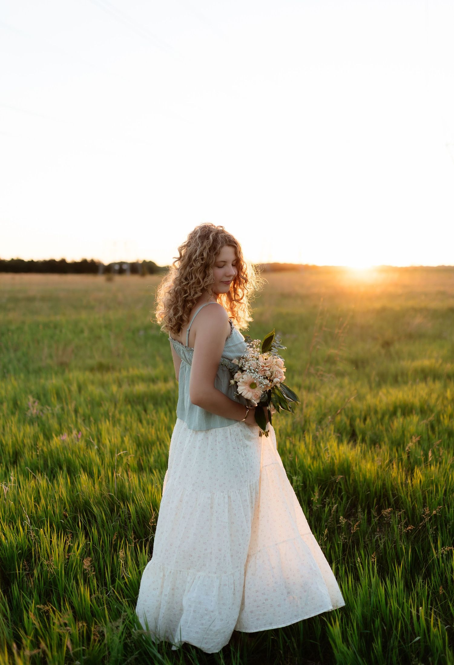 Young woman with curly hair holding floral bouquet standing in green field during golden sunset.