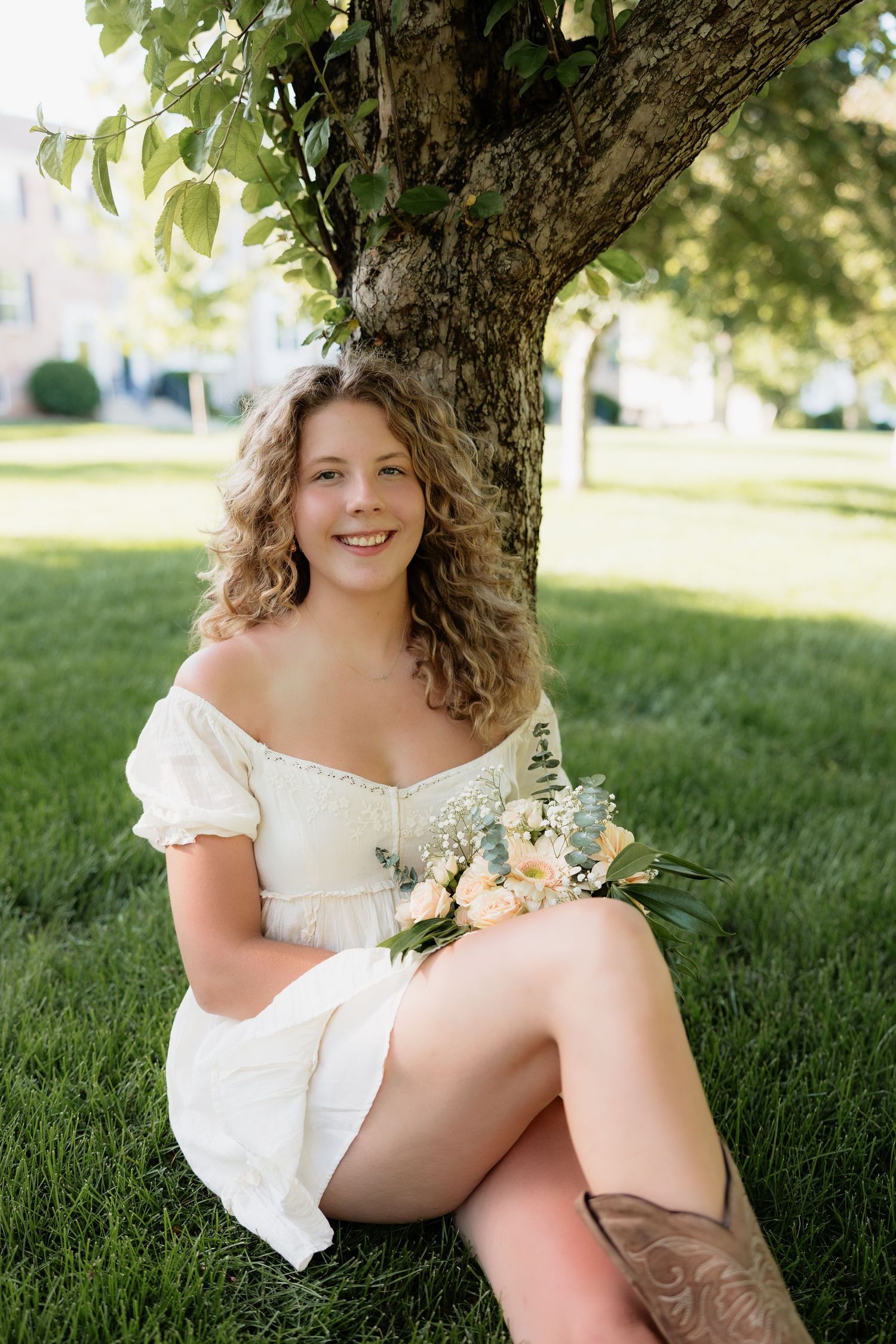 Smiling young woman in white off-shoulder dress holding a bouquet while sitting on grass under a tree.