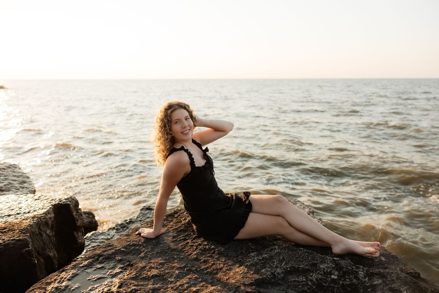 Woman in black dress posing on rocky shore by calm sea at sunset.