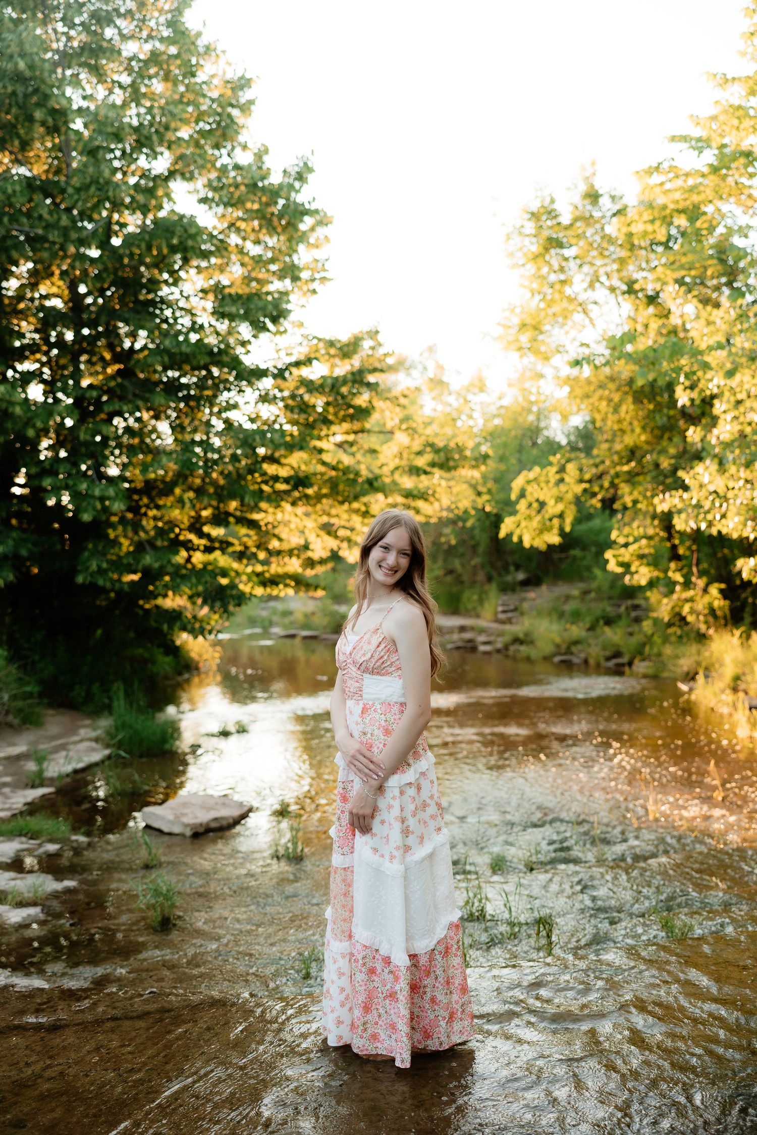 Pregnant woman in floral maxi dress standing in a sunlit creek surrounded by lush green trees.
