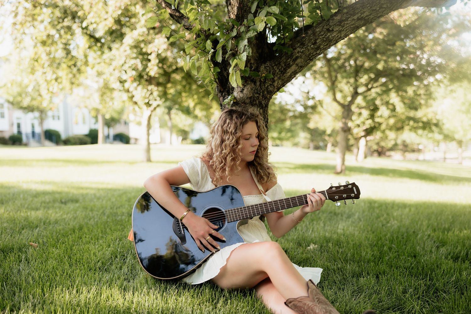 Young woman with curly hair playing acoustic guitar while sitting on green grass under a tree in a sunny park.