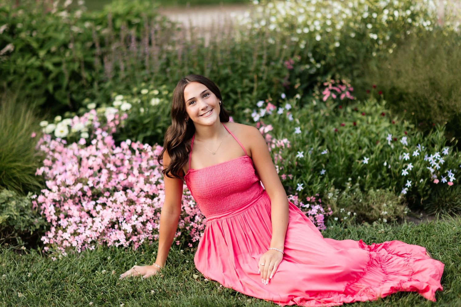 Young woman in coral dress sitting on grass surrounded by colorful garden flowers.