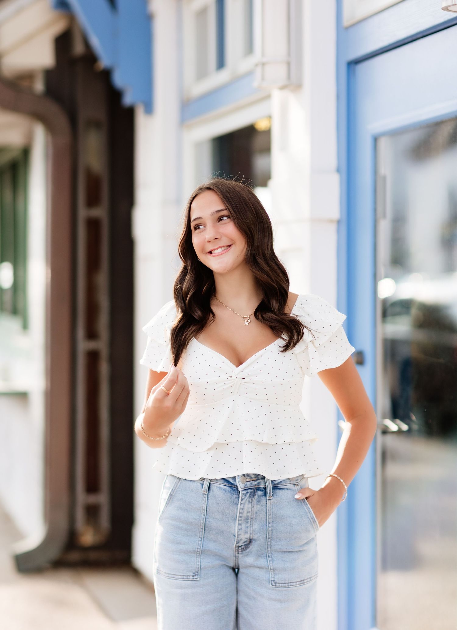 Young woman in white ruffled top and light jeans looking away smiling outside blue-trimmed storefront.