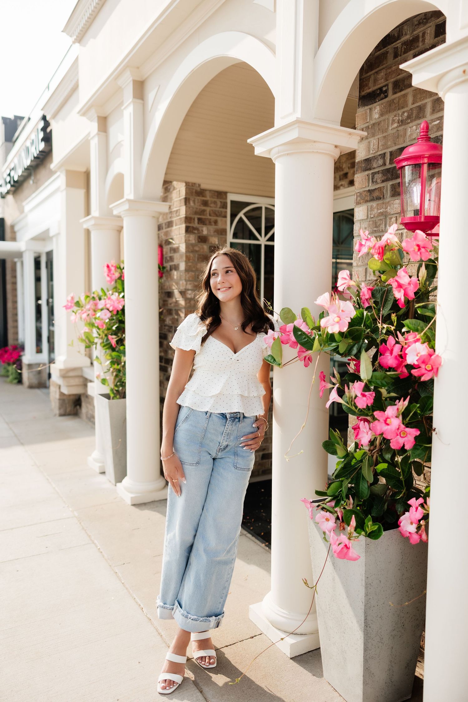 Young woman in white crop top and light jeans stands by white columns adorned with bright pink flowers.