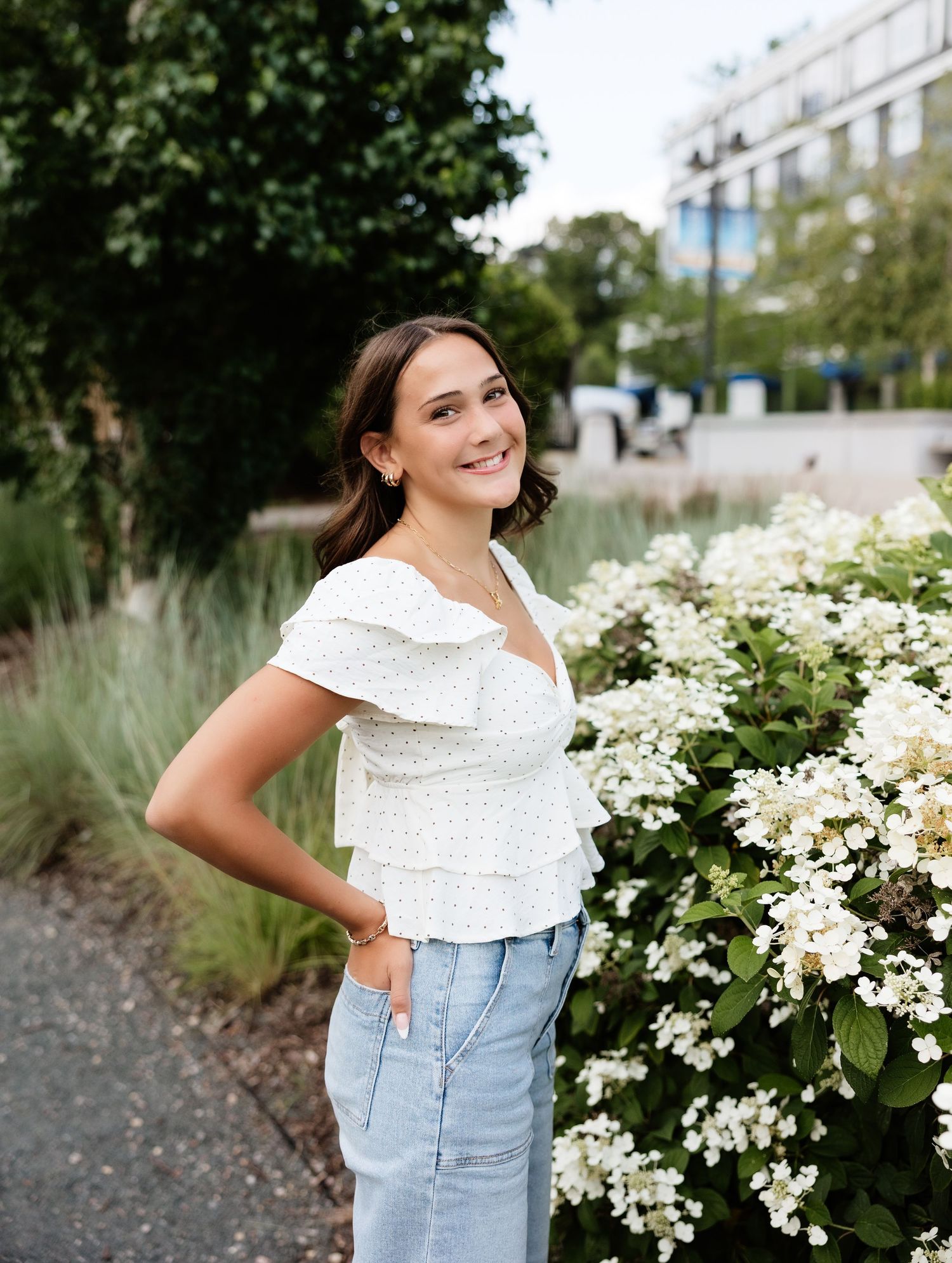 Young woman in white floral top and jeans smiling beside white hydrangeas in an urban garden setting.