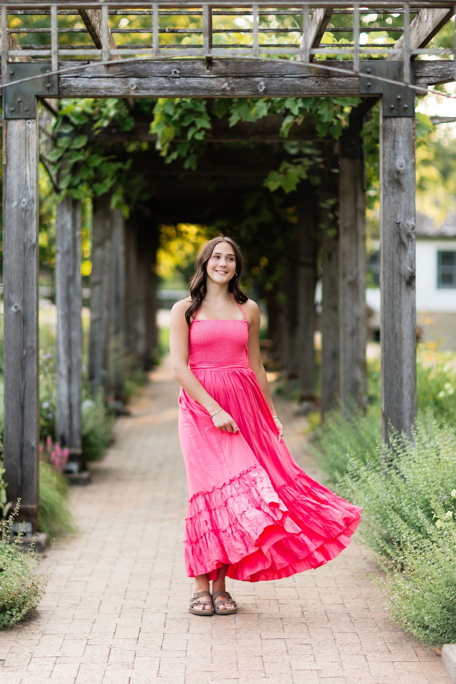 Smiling young woman twirling pink maxi dress under wooden pergola garden path.