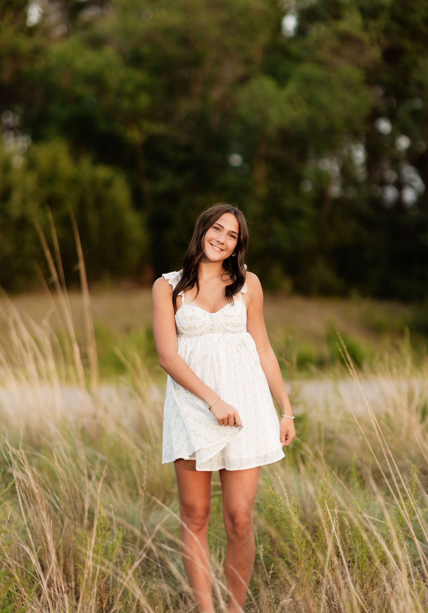 Smiling brunette woman in white sundress standing in tall golden grass field with green trees behind her.