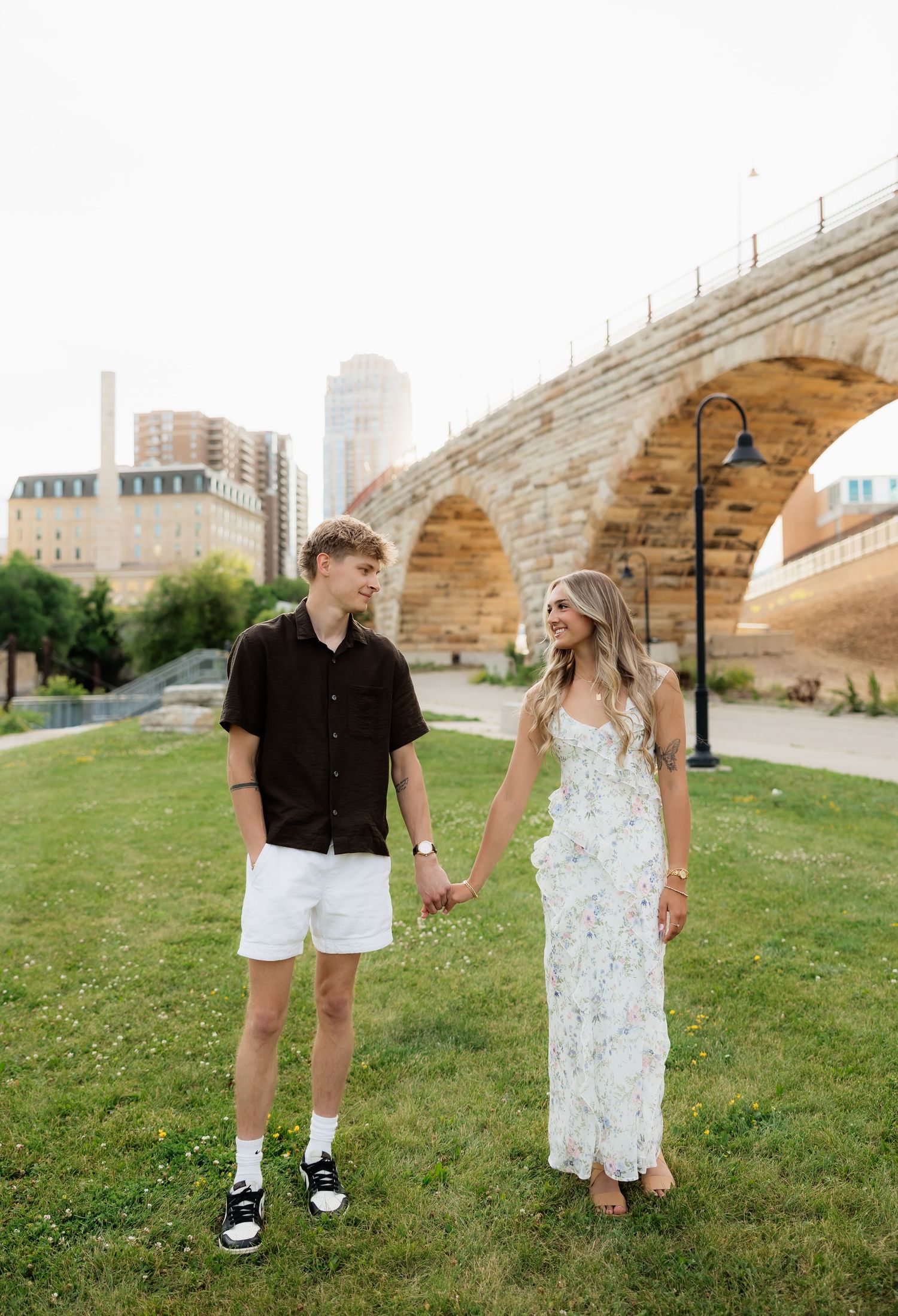Young couple holding hands on green grass in front of a historic stone arch bridge.