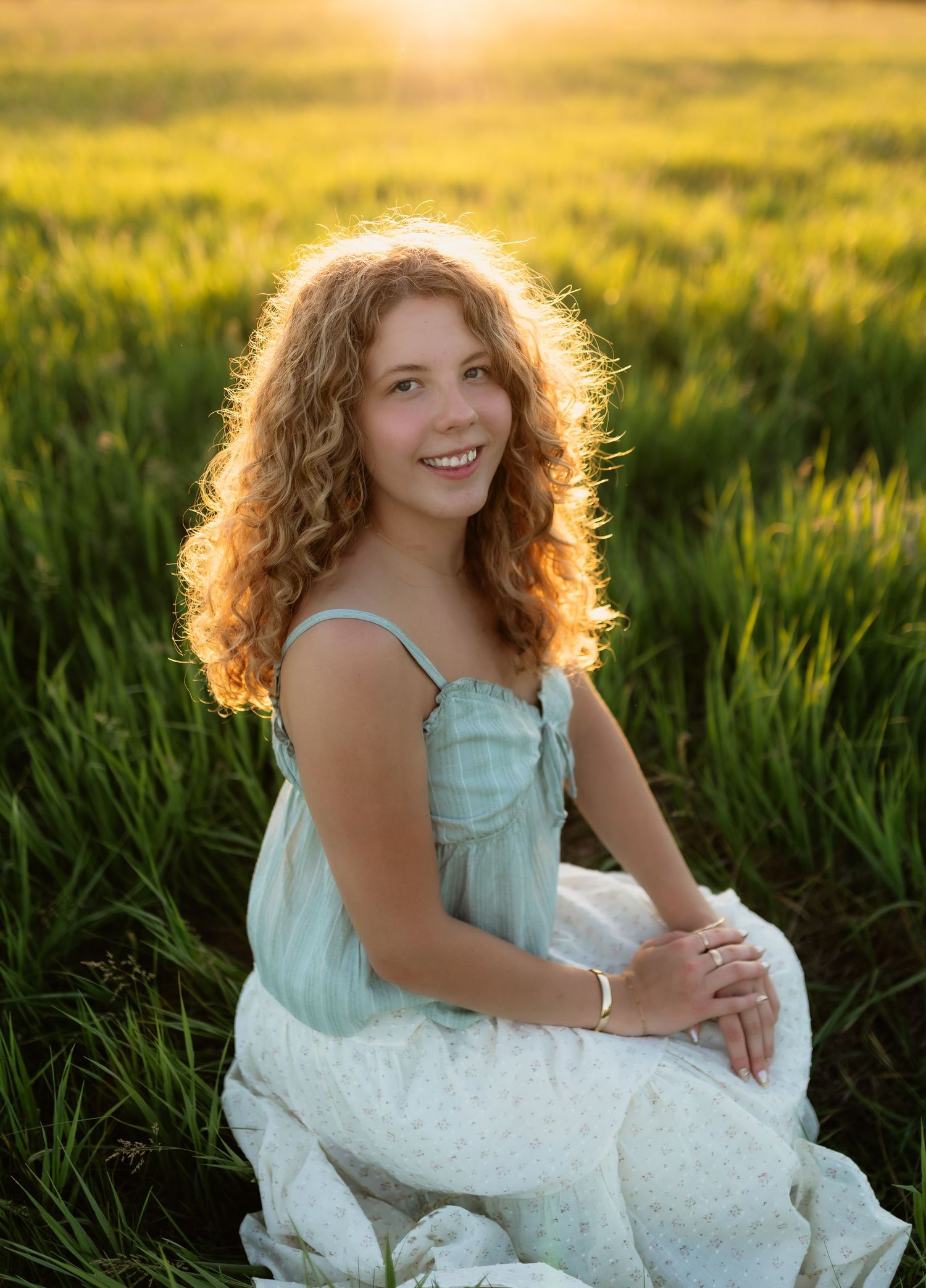 Smiling teenage girl with curly auburn hair in a mint dress sitting in a sunlit green field at golden hour.