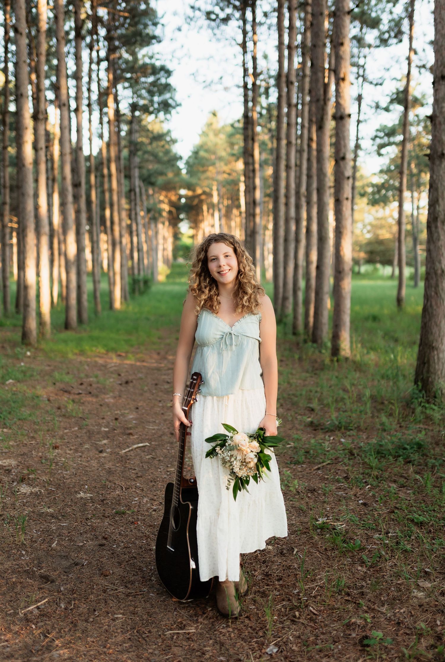 Smiling young woman with curly hair holding a guitar and flower bouquet while standing on a forest path at golden hour.