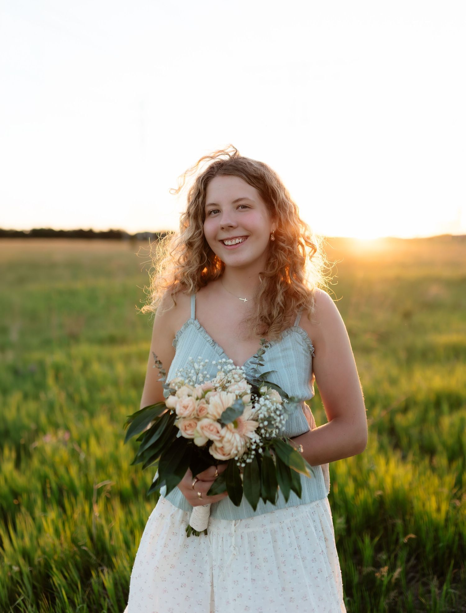 Young woman with curly hair holding white flowers, smiling outdoors in a field at sunset.