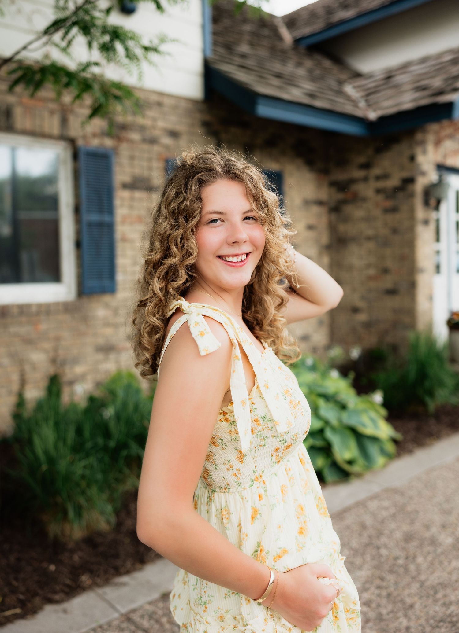 Smiling young woman with curly blonde hair in a yellow floral sundress posing outdoors near a brick house.