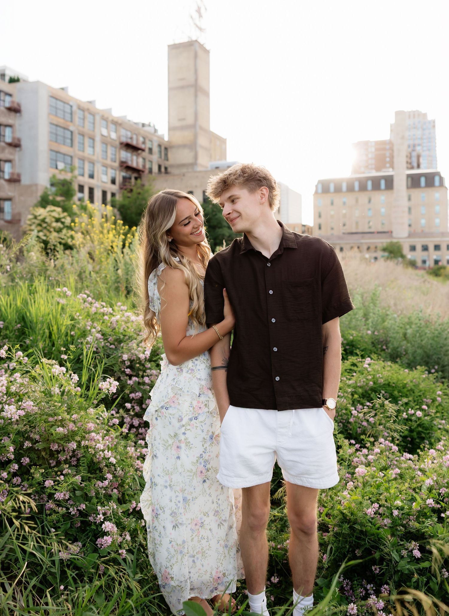 Smiling couple standing together in a lush urban wildflower garden with city buildings behind.