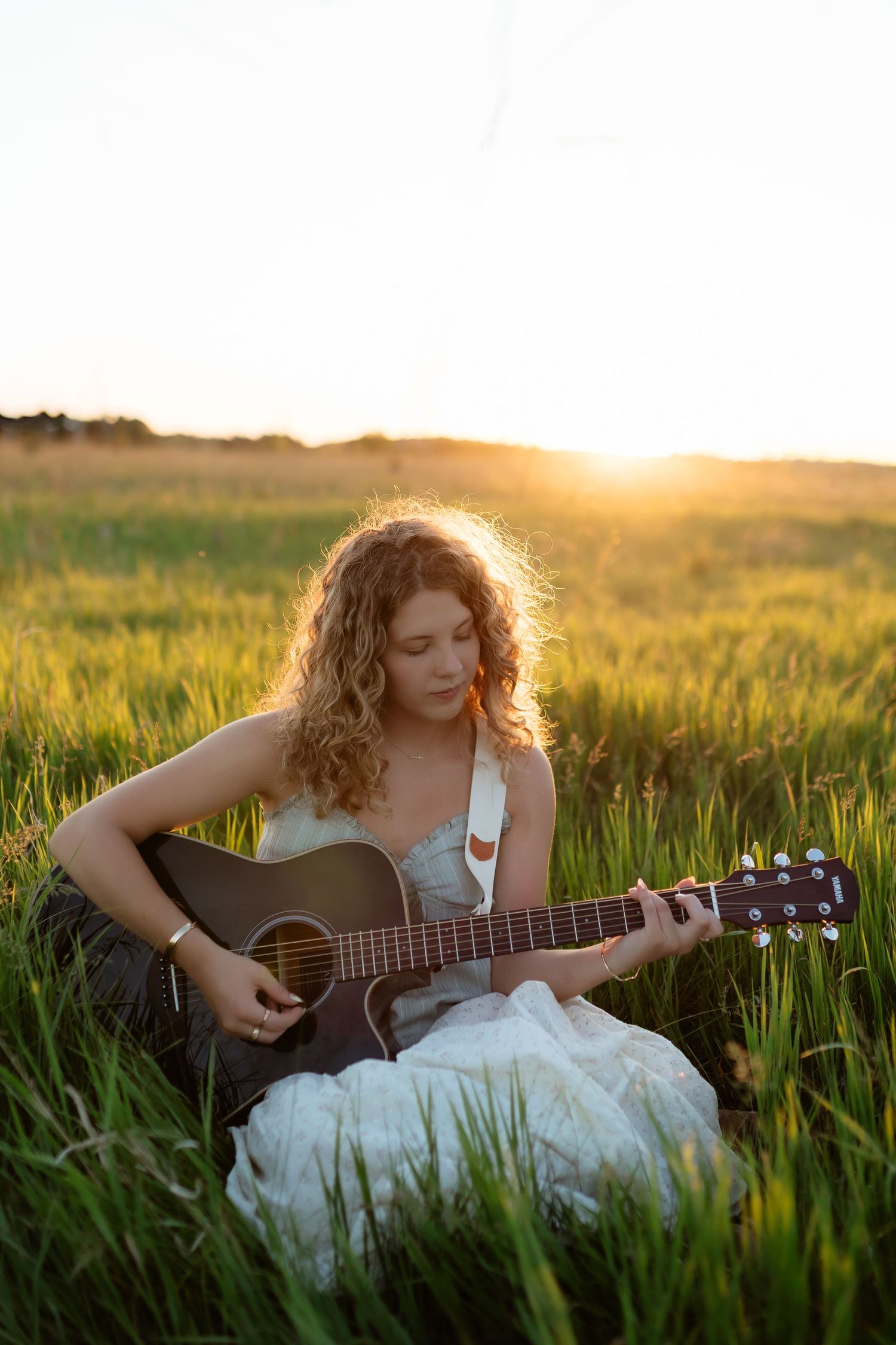Young woman with curly hair playing acoustic guitar while sitting in lush green field at sunset.