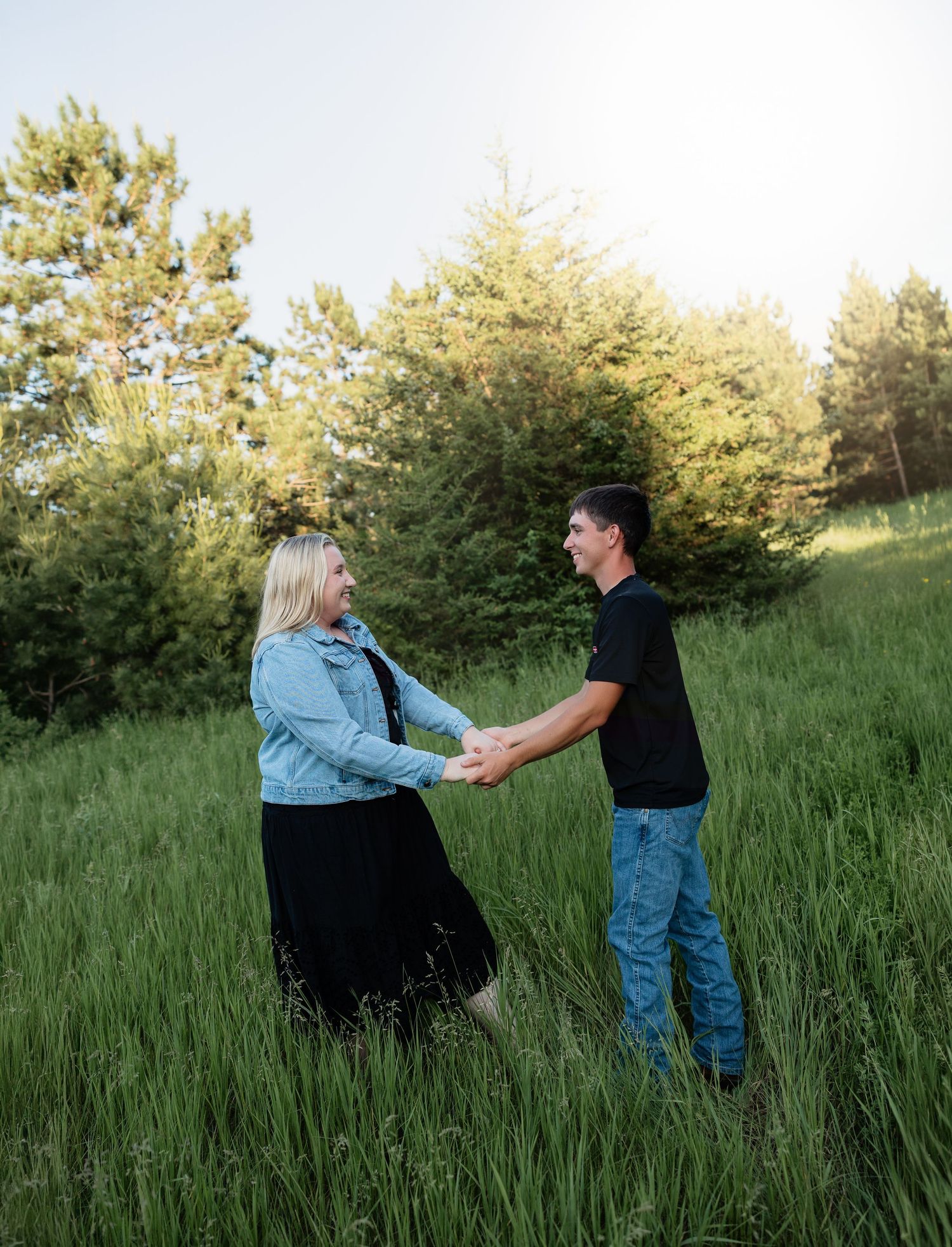 Couple holding hands and smiling in a lush green meadow, woman in denim jacket and black skirt, man in black shirt and jeans.