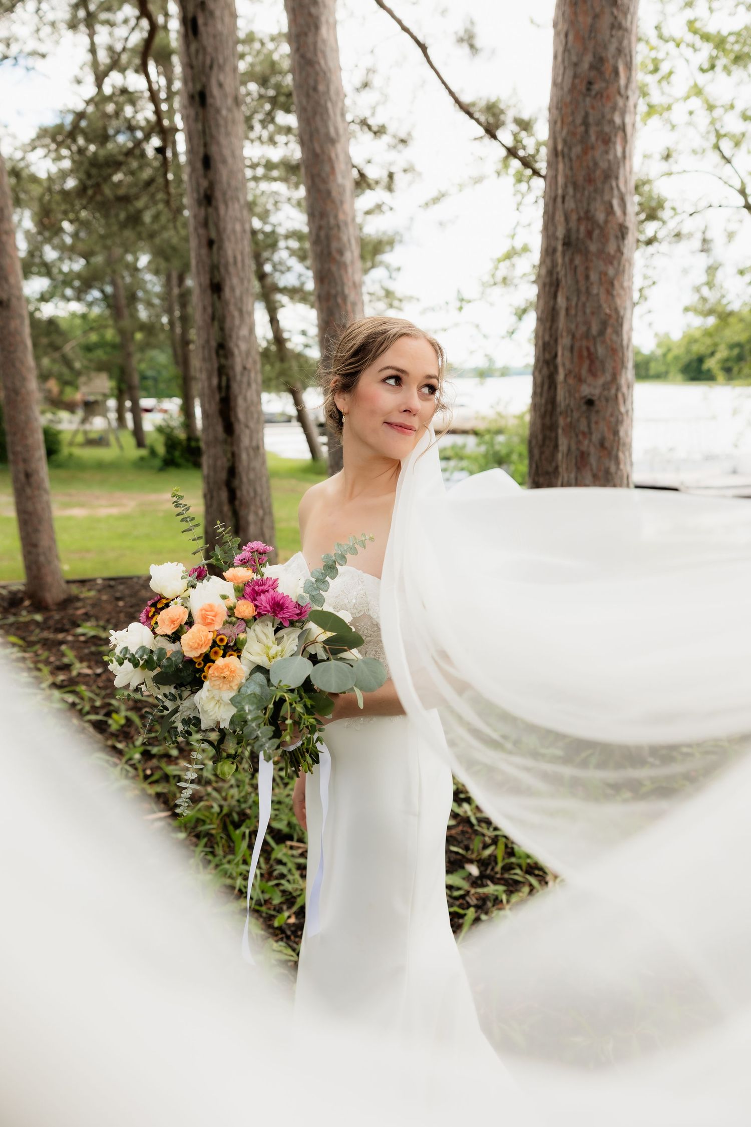 Bride in white gown holding colorful bouquet with veil flowing in breeze among tall trees near lakeside.
