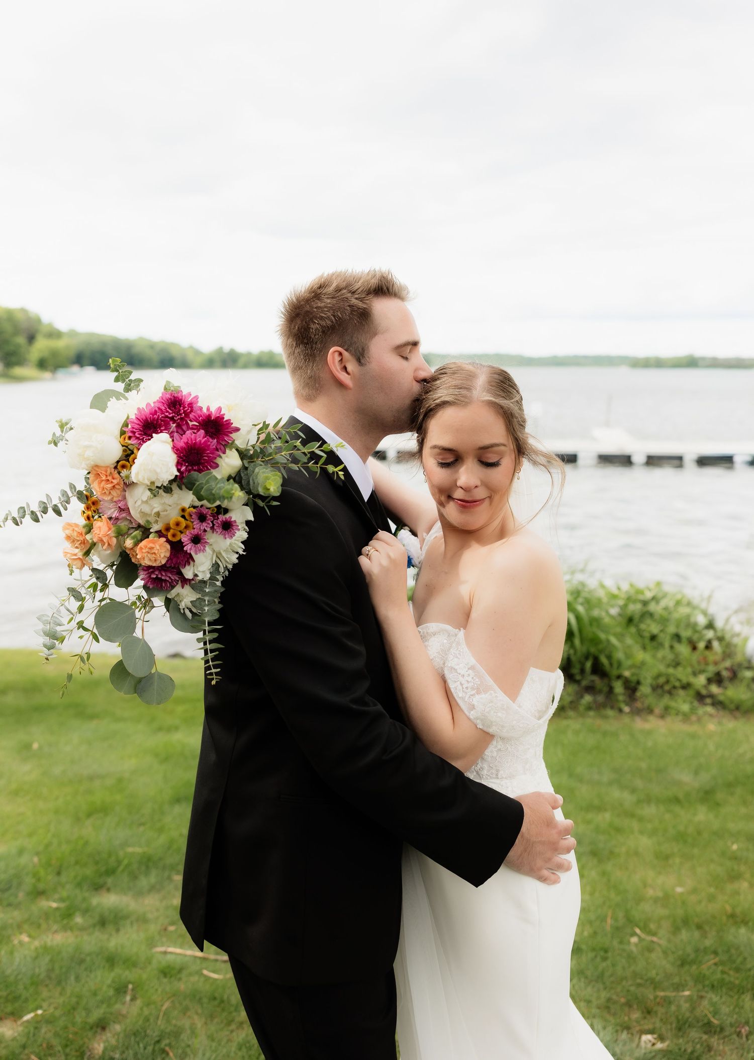 Groom in black tuxedo kisses bride holding colorful bouquet by lakeside on their wedding day.