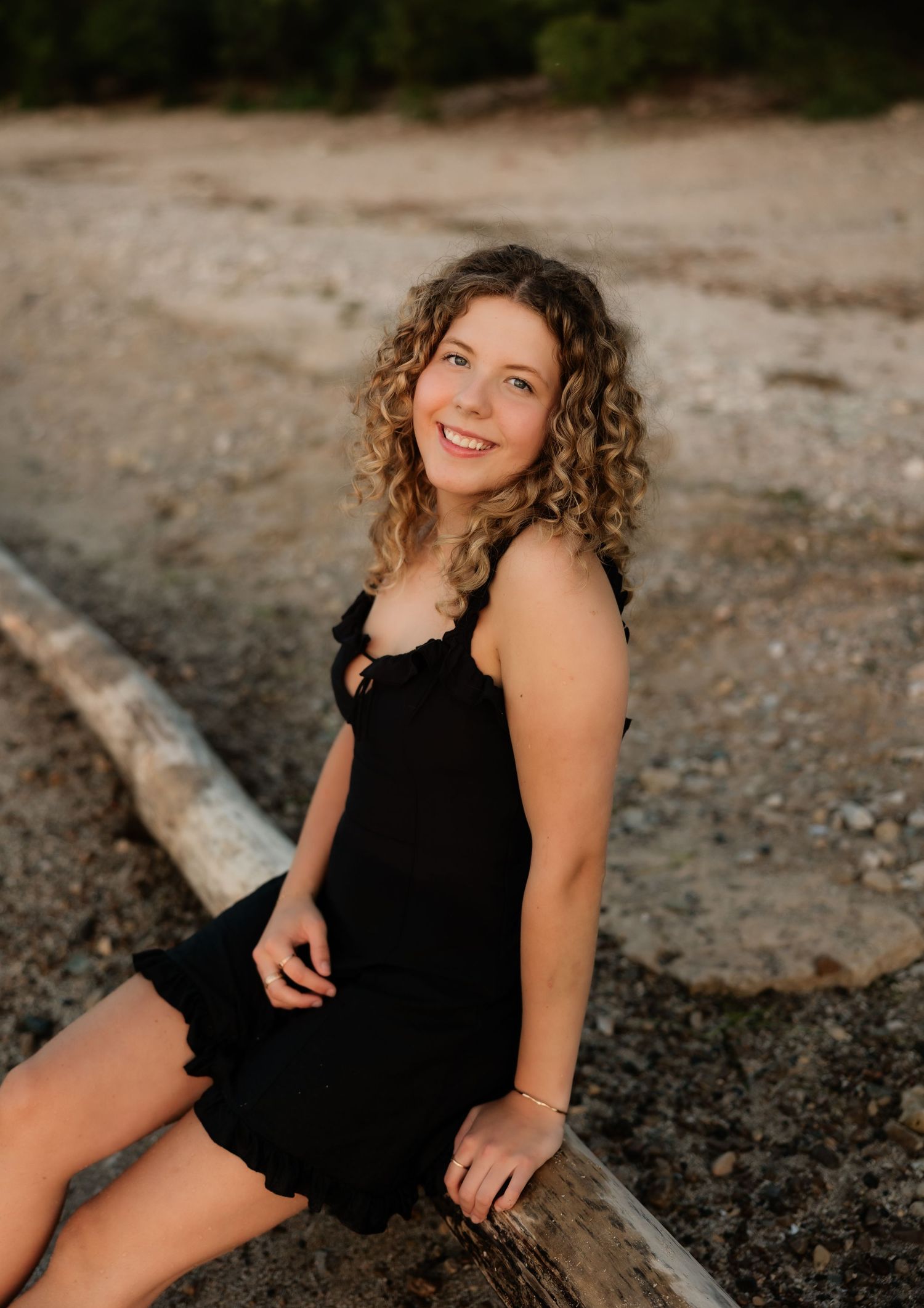 Smiling young woman with curly hair in black dress leaning on driftwood on a rocky beach.