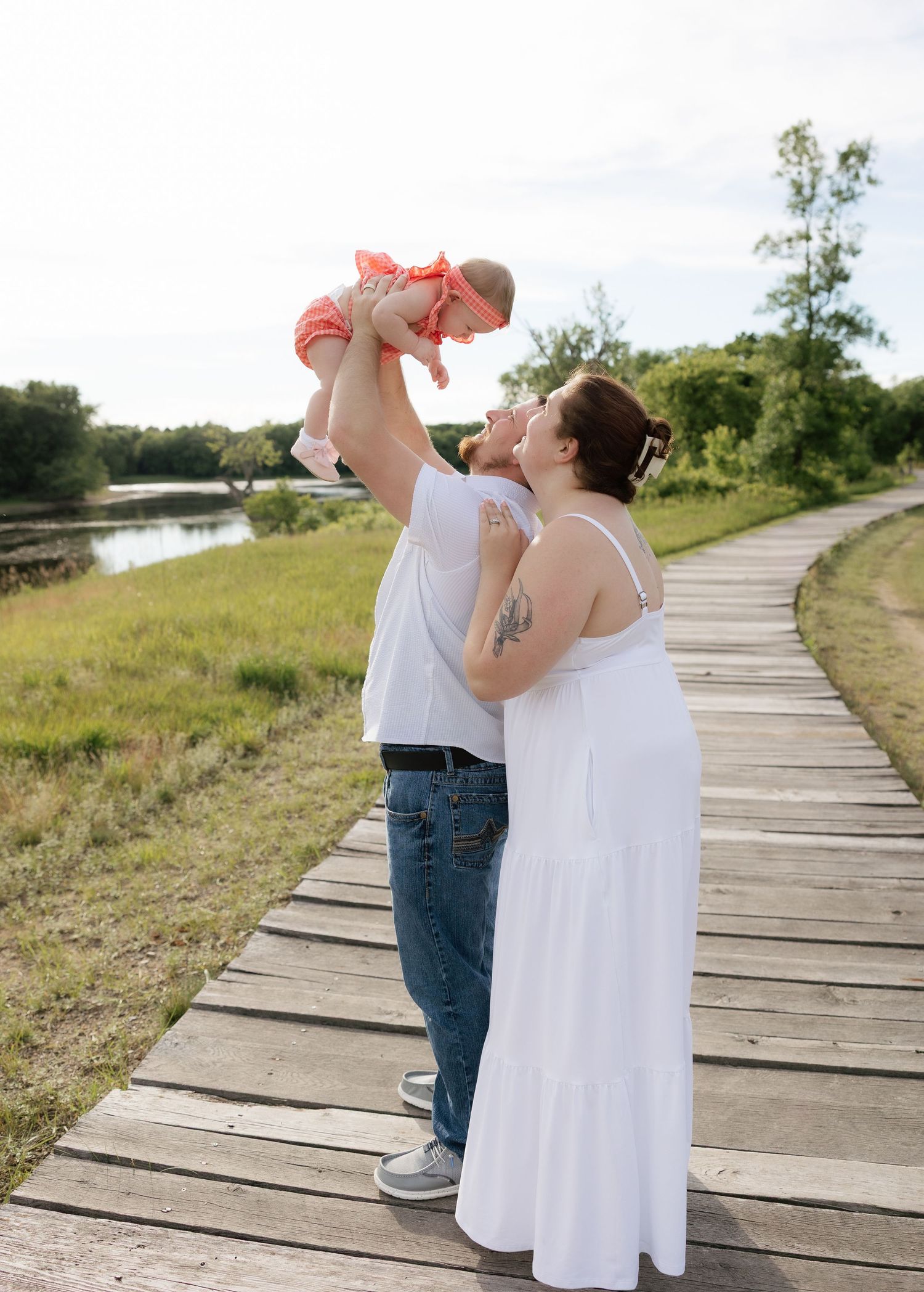 A couple stands on a wooden boardwalk, man lifting baby in pink dress while woman in white gown looks on, near a scenic river.