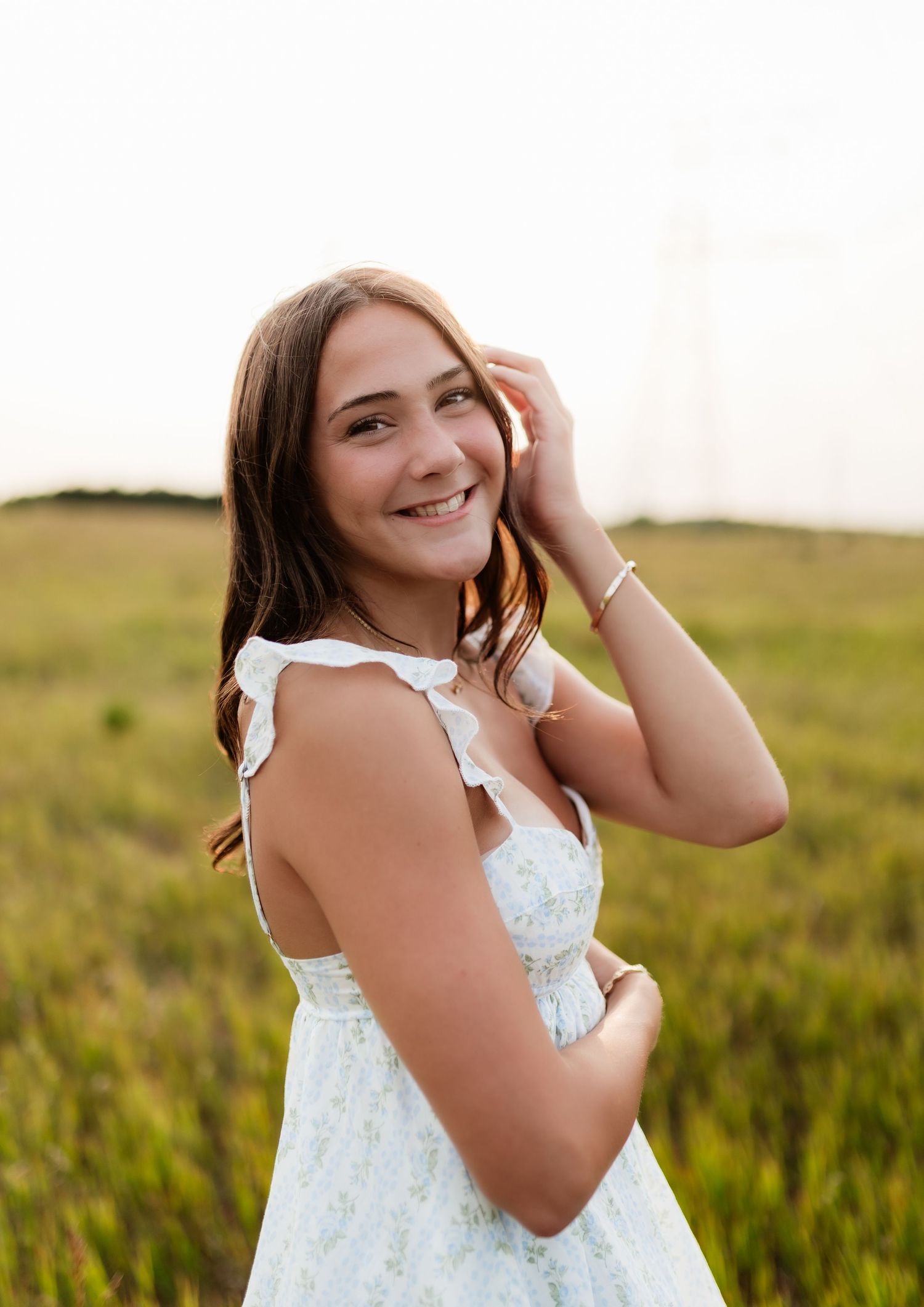 Young woman in white floral dress smiling in golden sunlit grassy field.