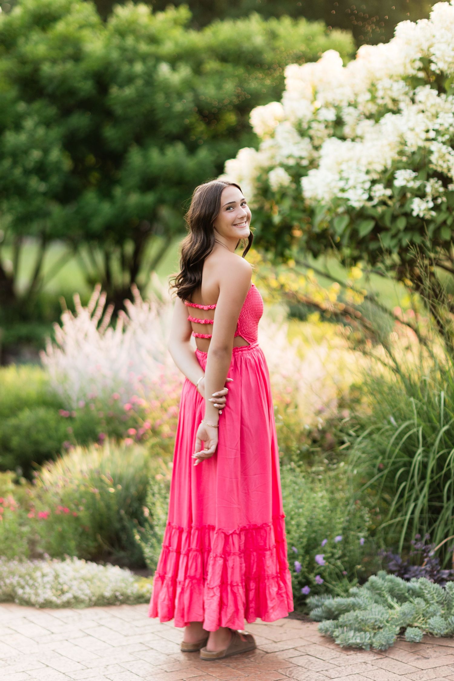 Young woman in hot pink ruffled maxi dress posing from behind in a colorful garden setting.