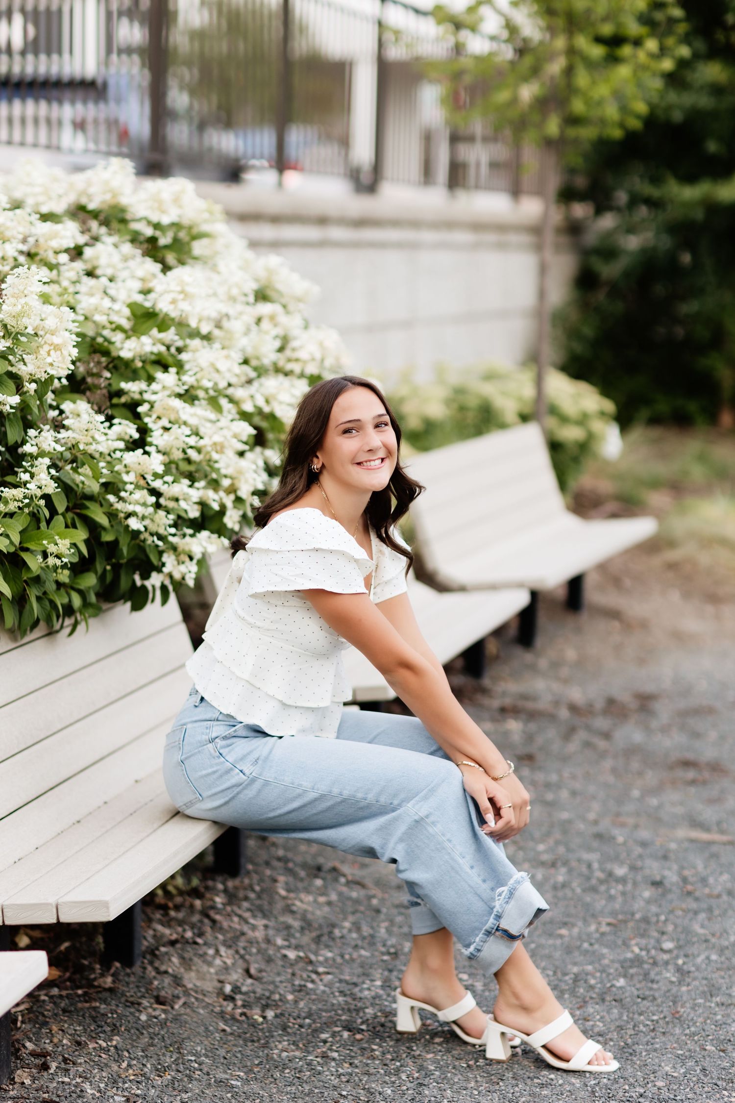 Young woman in white top and jeans sitting on a bench beside white hydrangeas, smiling outdoors.