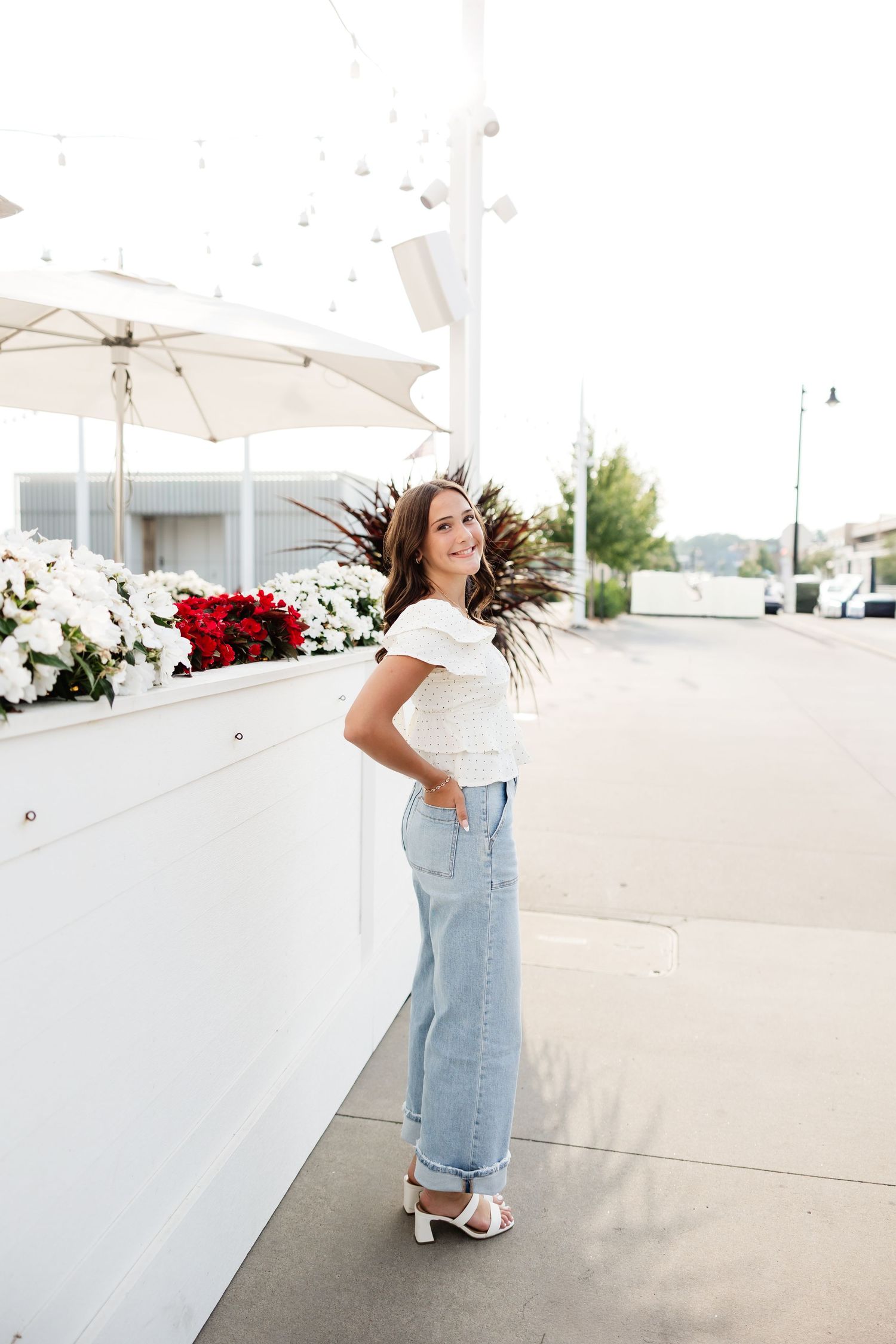 Brunette woman in white off-shoulder top and jeans posing beside outdoor flower planters on sidewalk.
