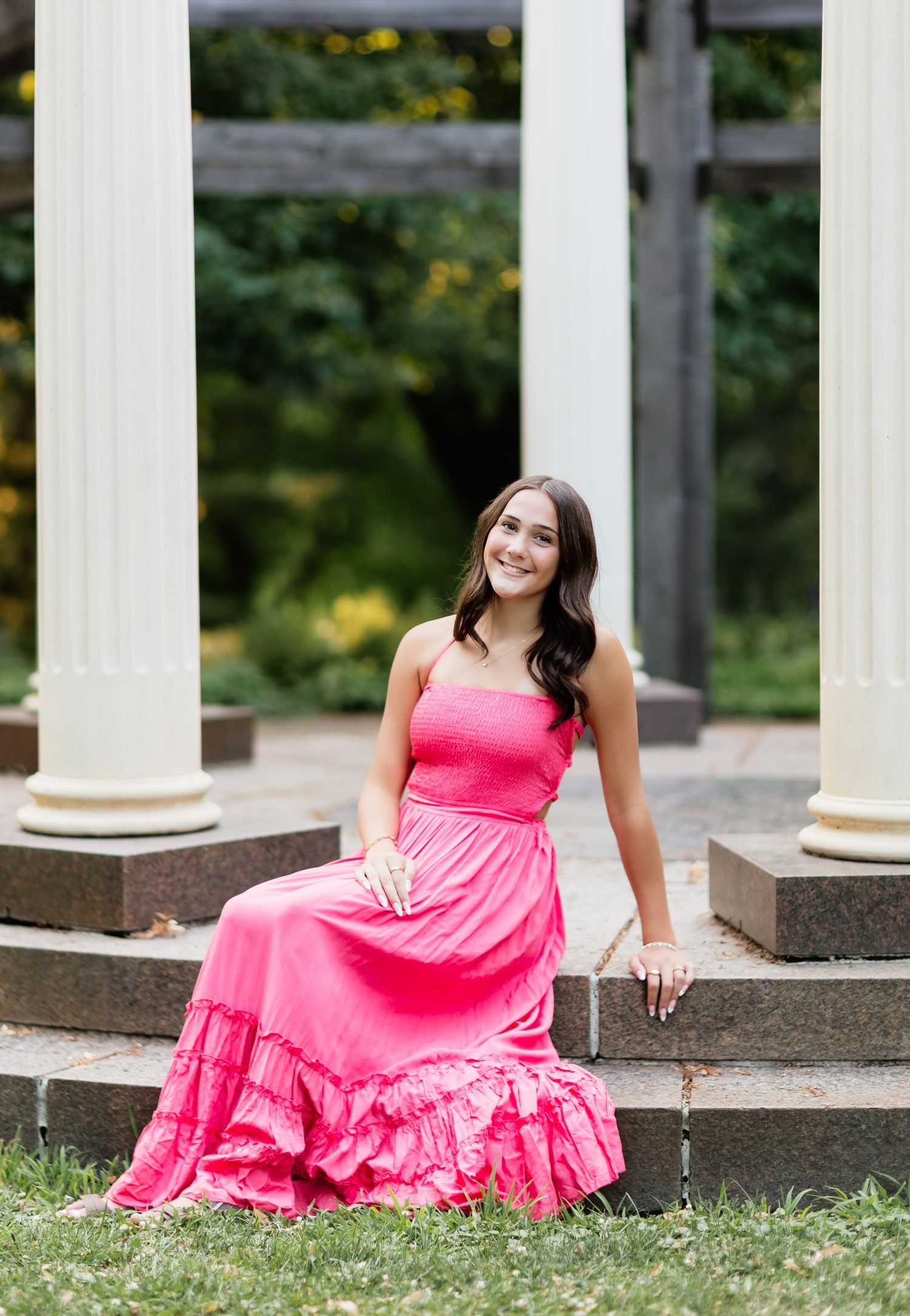 Smiling young woman in pink maxi dress seated on steps near white columns.