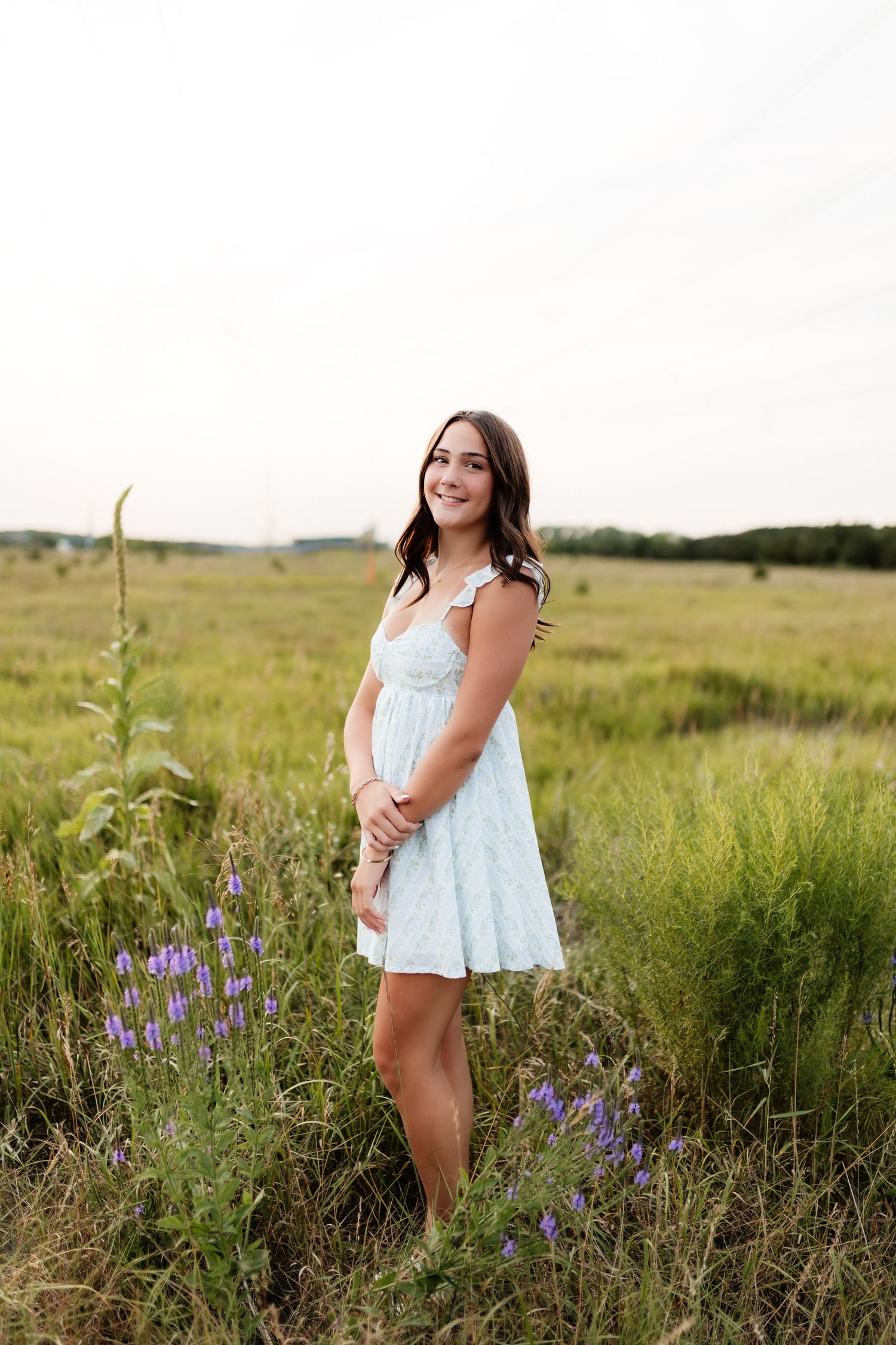 Smiling young woman in a light blue sundress standing in a wildflower meadow with purple blooms.